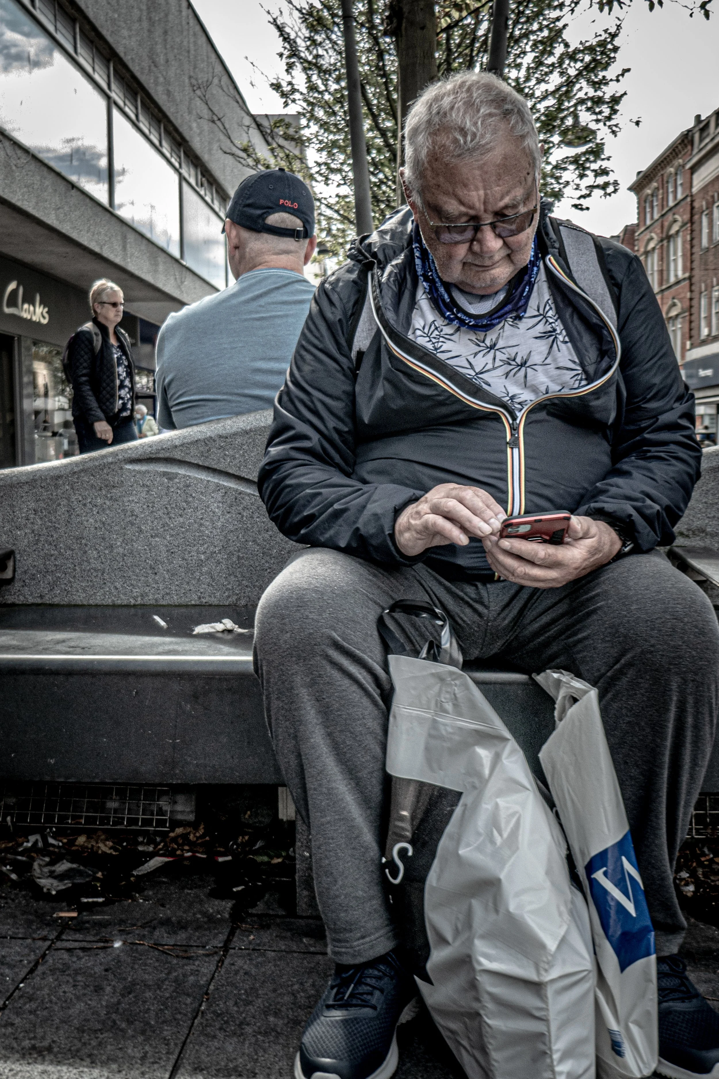 An elderly man sitting on a bench, looking down at his phone, with a shopping bag beside him, in an urban outdoor setting with pedestrians and buildings in the background.