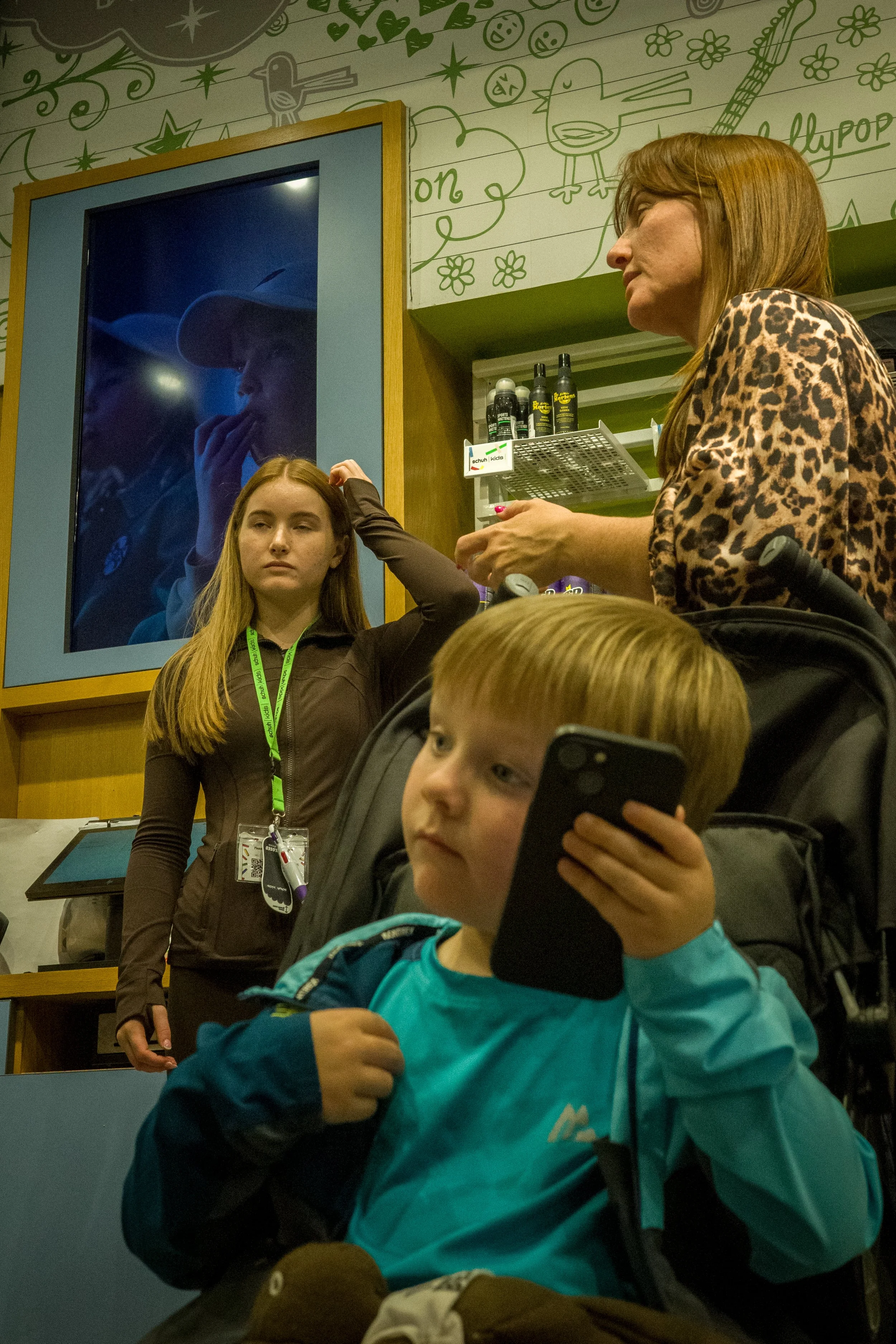 A woman with long red hair and a leopard print shirt talking to a young boy with blond hair holding a smartphone. In the background, a large mirror reflecting a woman in a cowboy hat and a blue jacket. Children are in the foreground, one with a backp