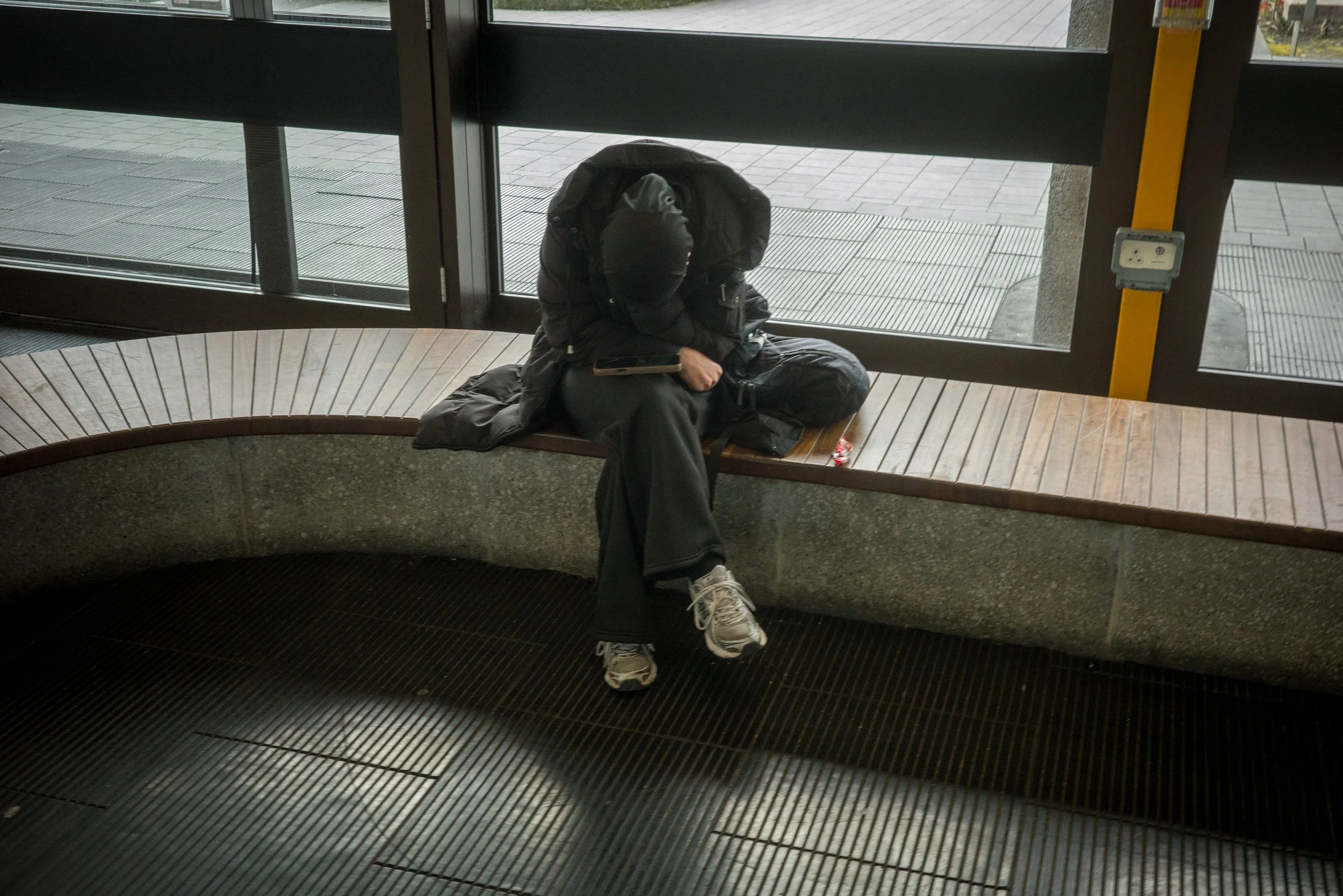 A person sitting on a curved wooden bench inside a building near glass doors. The person is hunched over with their head down, wearing a black beanie, black jacket, and gray pants. They are holding a phone in their right hand, with a backpack at thei
