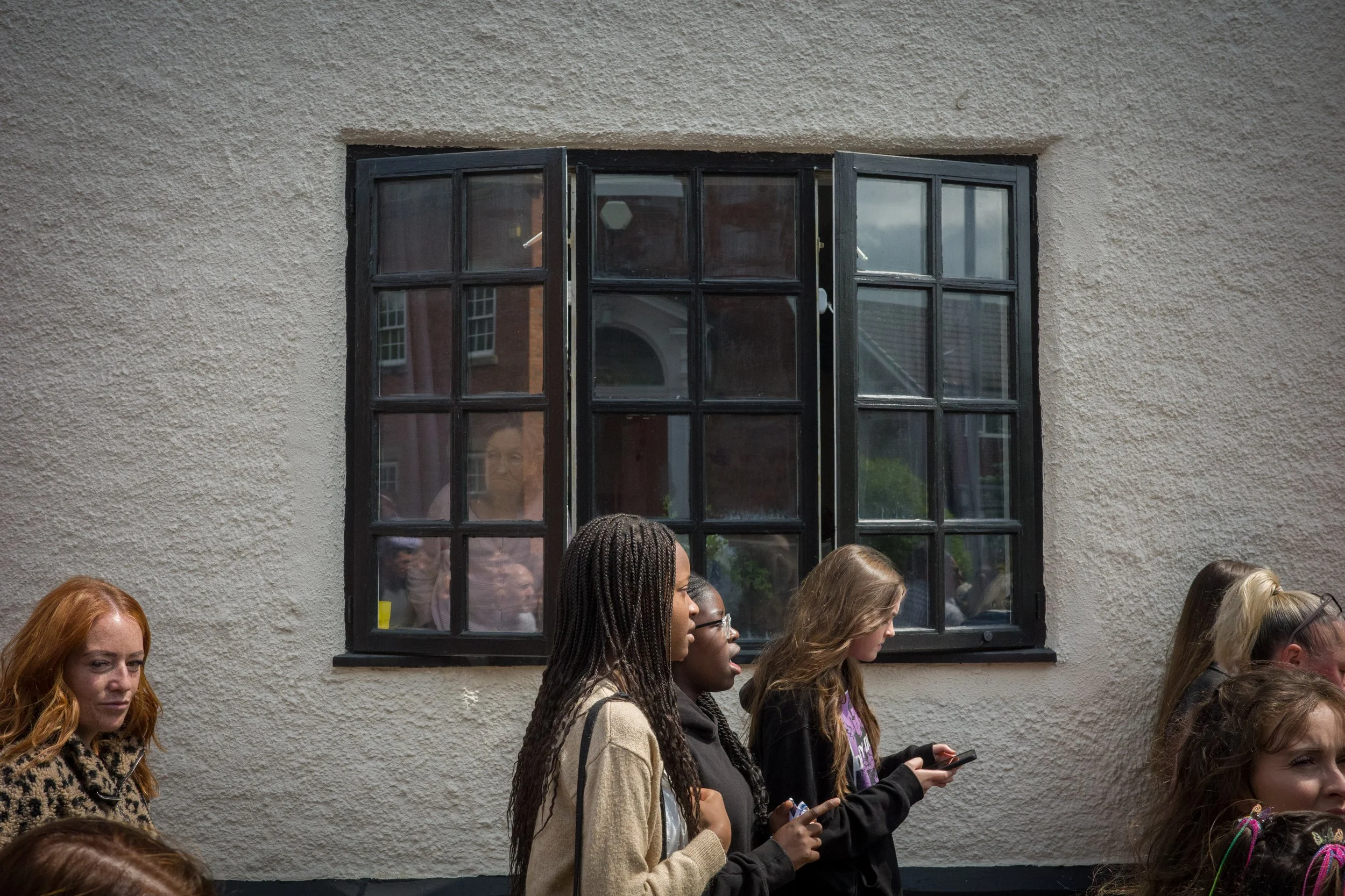 A group of people, mostly women, standing outdoors in front of a beige wall with a black-framed window. Some are looking down at their phones, others are looking ahead. One woman has red hair, and others have different hair colors and styles. A refle