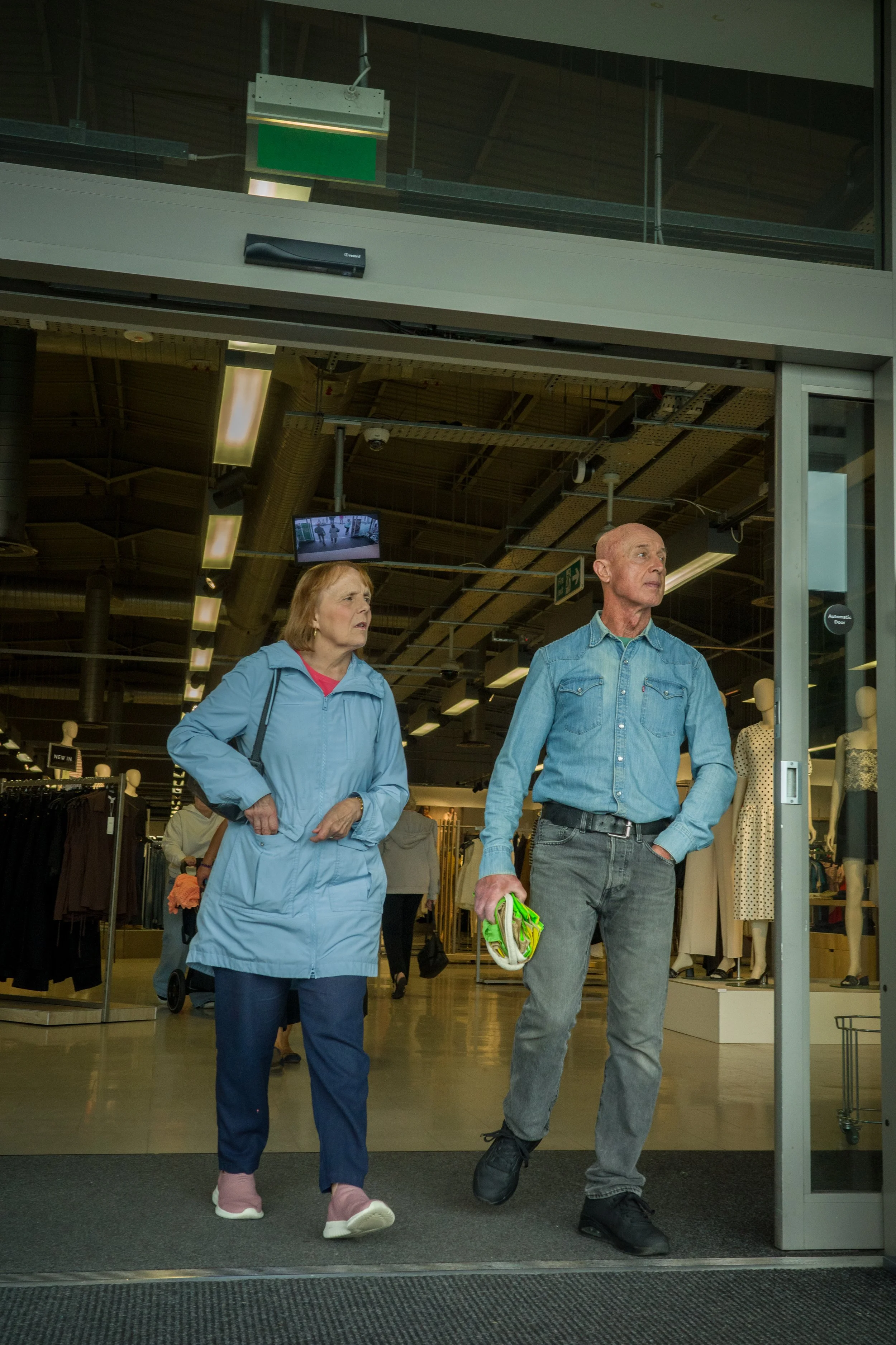 An older man and woman are walking out of a clothing store, looking to the side. The man is holding a green and white item, dressed in a denim shirt and gray jeans. The woman is wearing a light blue coat and dark pants. The store has mannequins and c