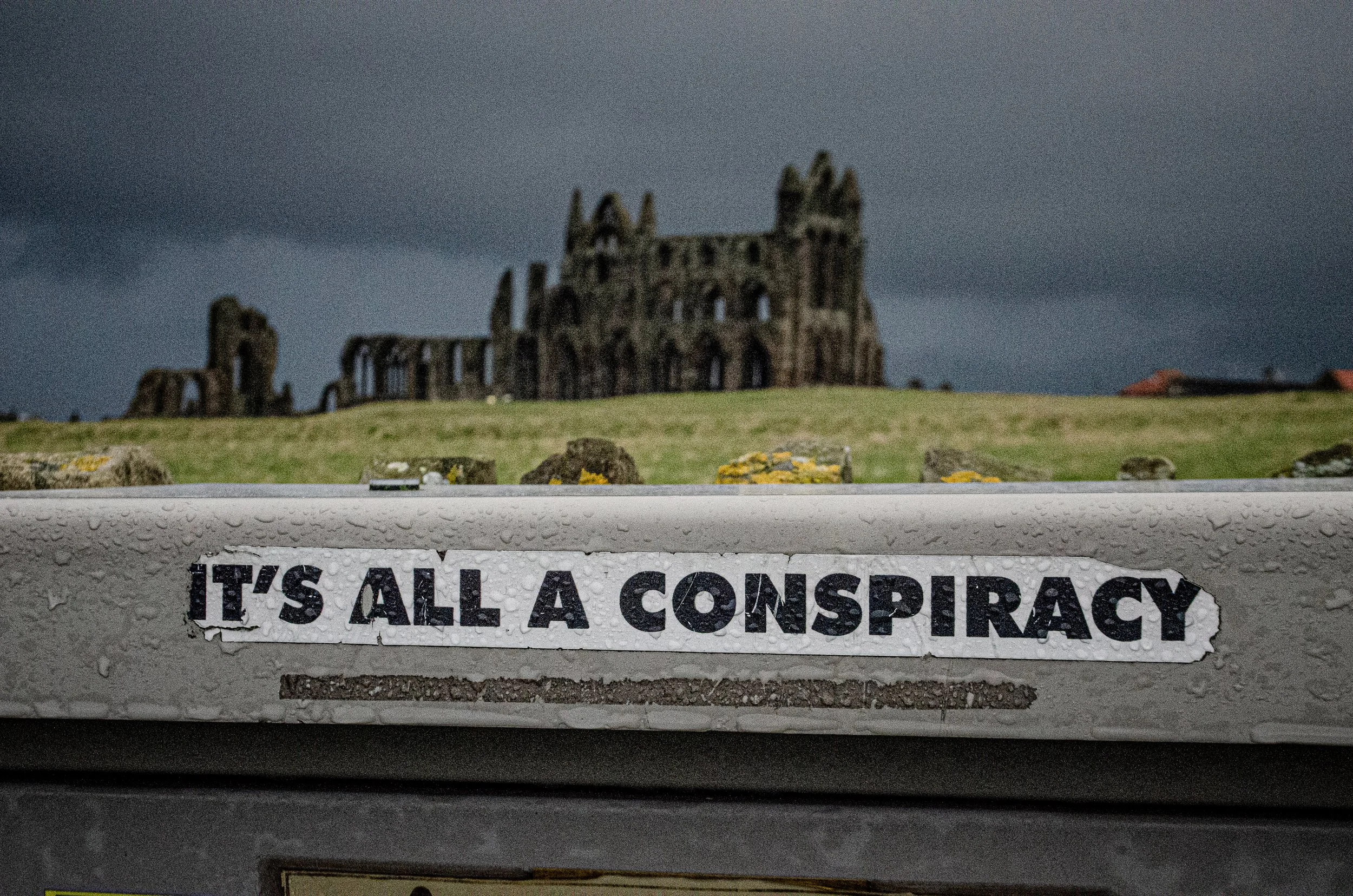 A sticker on a surface with the text 'IT'S ALL A CONSPIRACY'. In the background, there is a ruined castle on a hill under a dark cloudy sky, and the surface with the sticker is wet with raindrops.