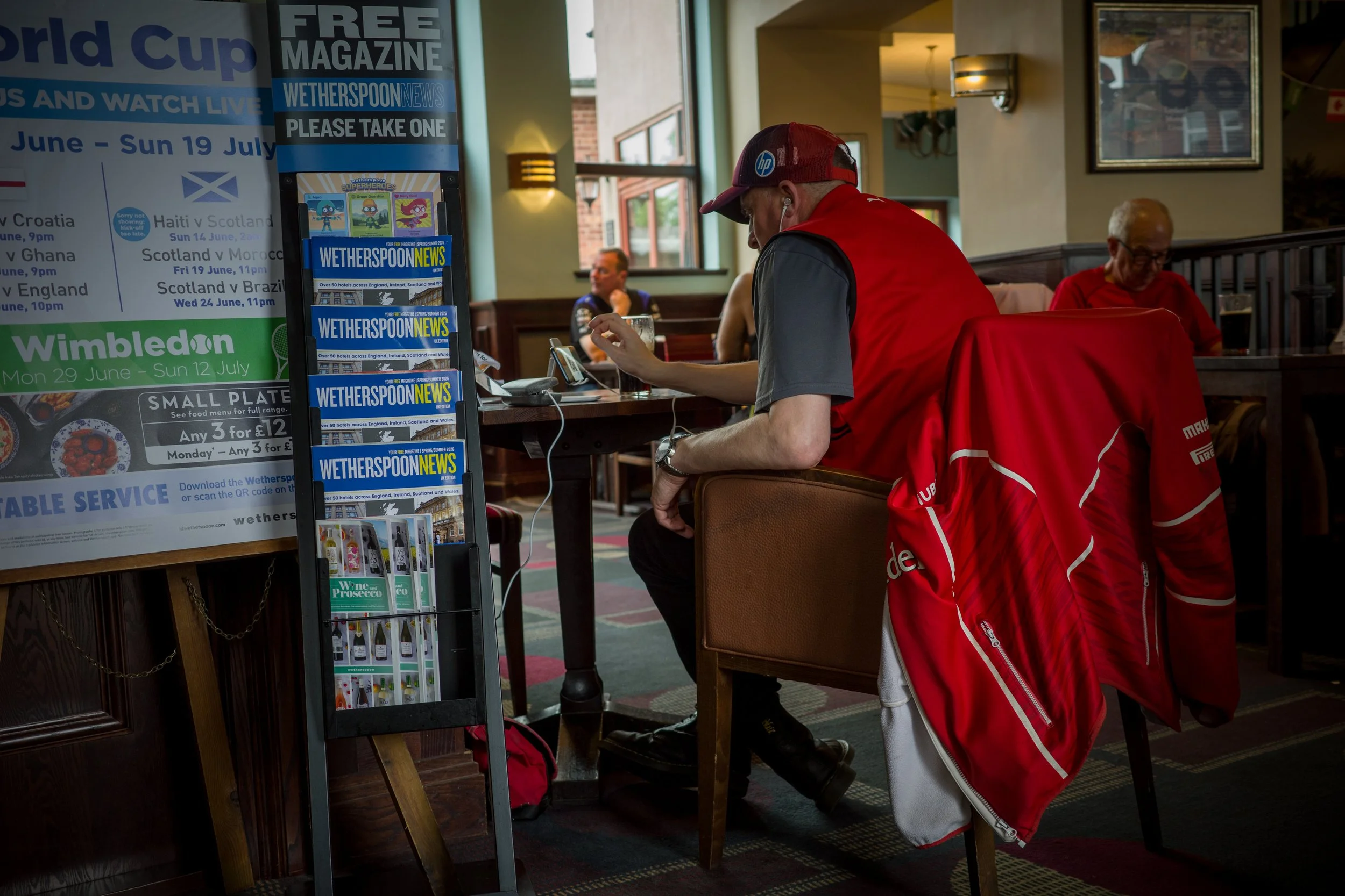 A man wearing a red cap and red jacket is sitting at a table using his phone inside a restaurant. There are other people sitting in the background, and a stand with brochures and magazines is visible in the foreground.