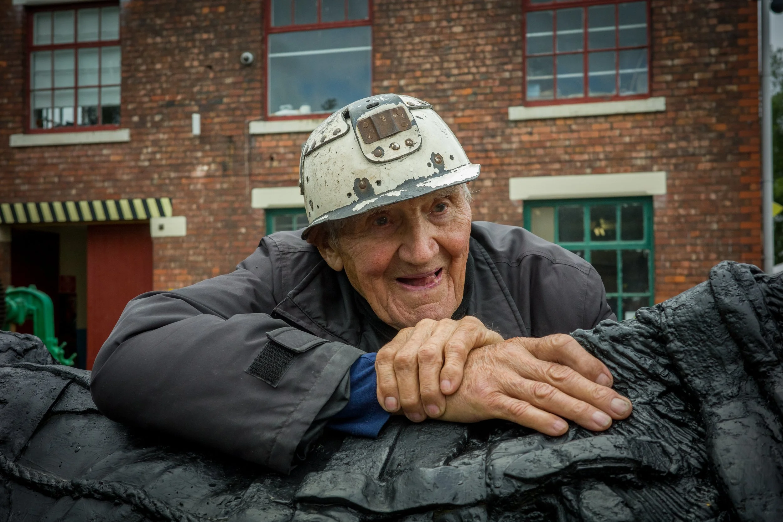 An elderly man wearing a white construction helmet, leaning on a large, black, textured surface outdoors, with a brick building in the background.