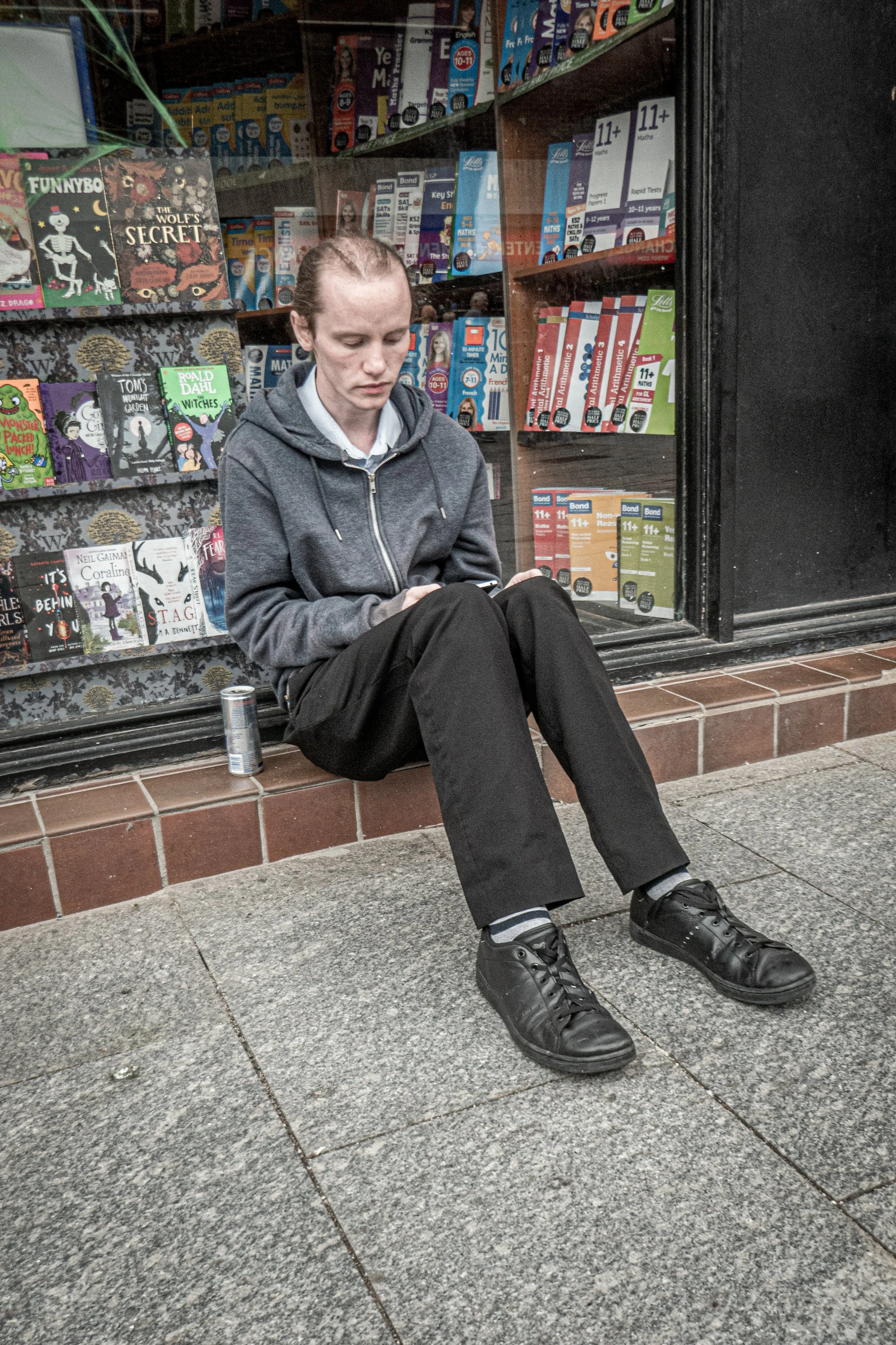 A young person with red hair, wearing a grey hoodie, black pants, and black shoes, sitting on a brick ledge outside a store with books displayed in the window, looking at their phone.