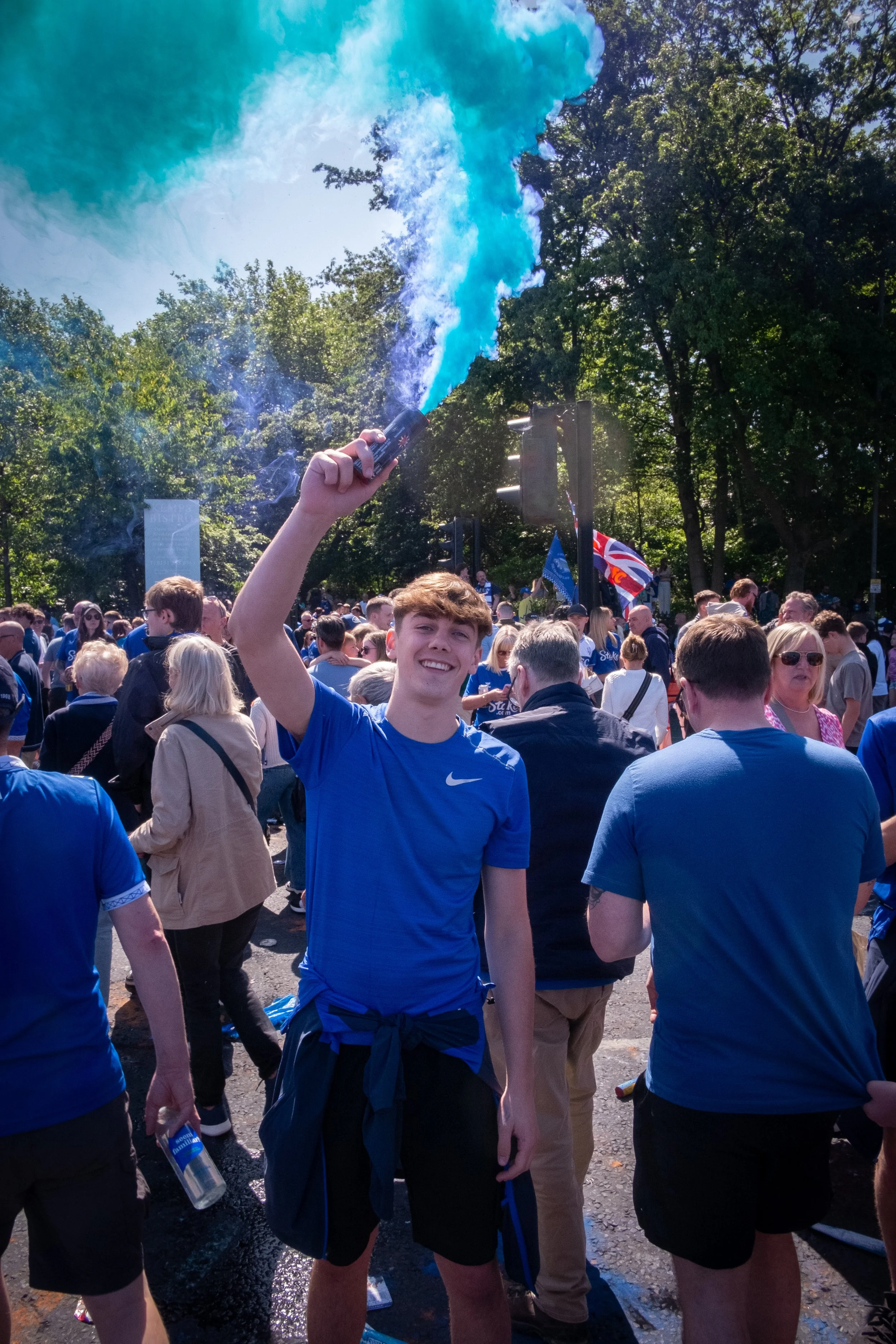 Young man smiling and holding a blue smoke flare at a crowd gathering, many dressed in blue, outdoors on a sunny day.