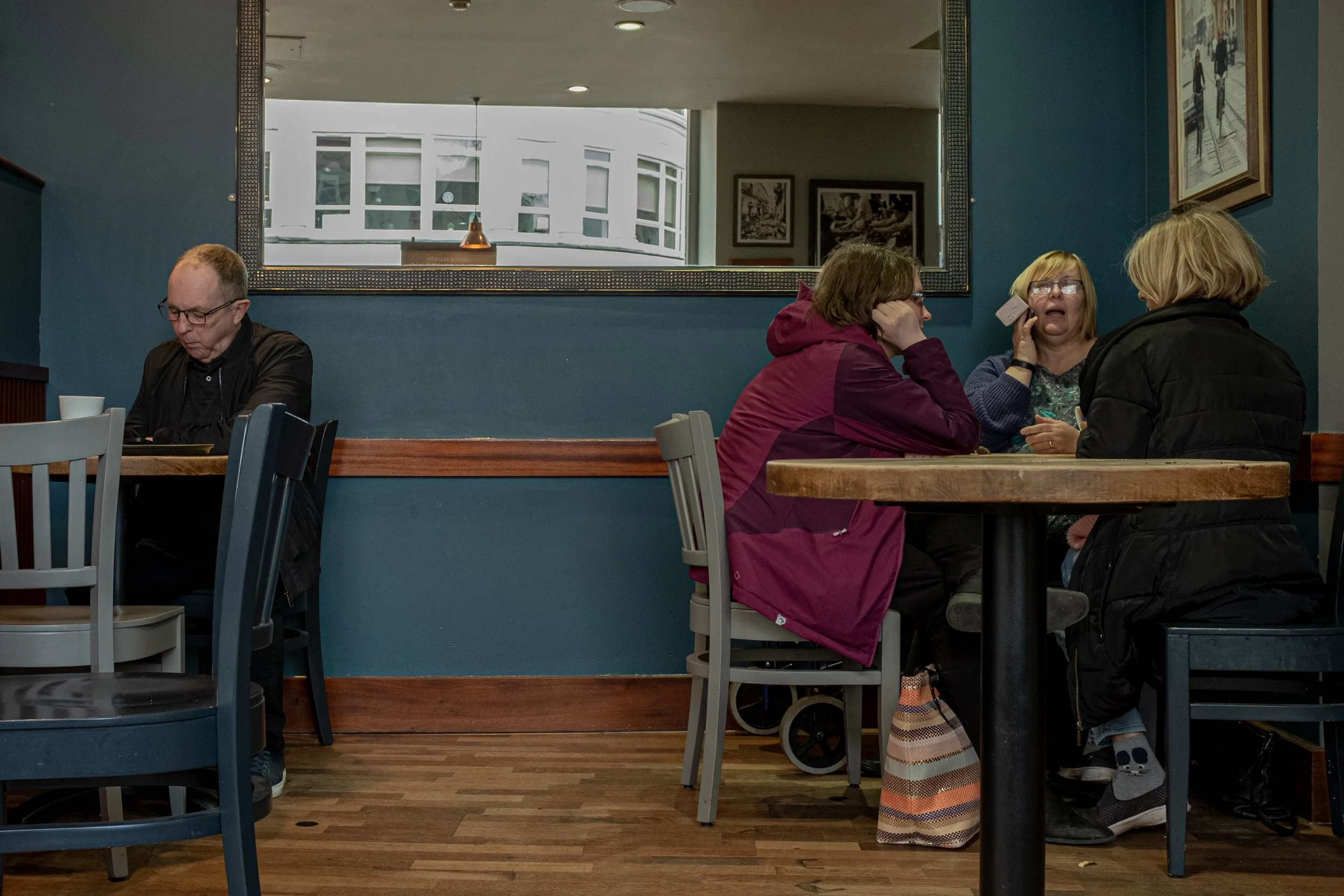 Three women and one man sit at tables in a cafe, with wallpaper, framed photos, and a large mirror on the walls, and windows showing a white building outside.