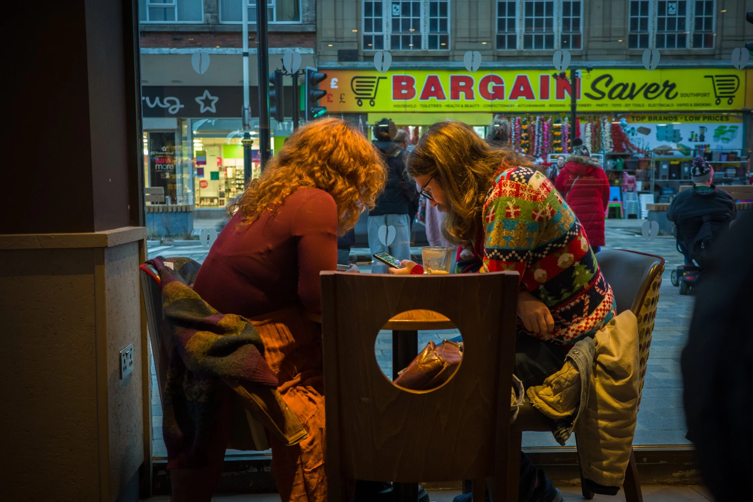 Two women sitting at a table near a large window in a cafe, looking at a phone, with an urban street scene outside featuring people and storefronts.