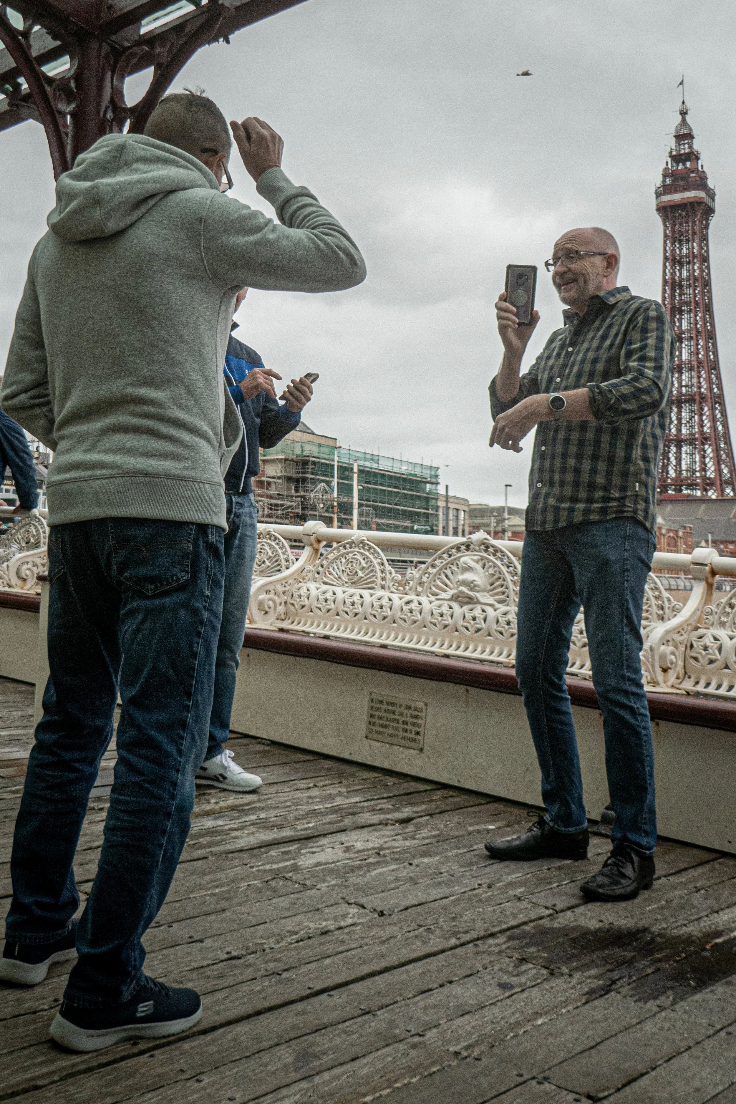 Four men standing on a wooden boardwalk near the Eiffel Tower in Paris, with a cloudy sky and a helicopter flying overhead. One man is wearing a hoodie, another is in a blue jacket, a third in a plaid shirt, and the fourth is in a dark jacket using a