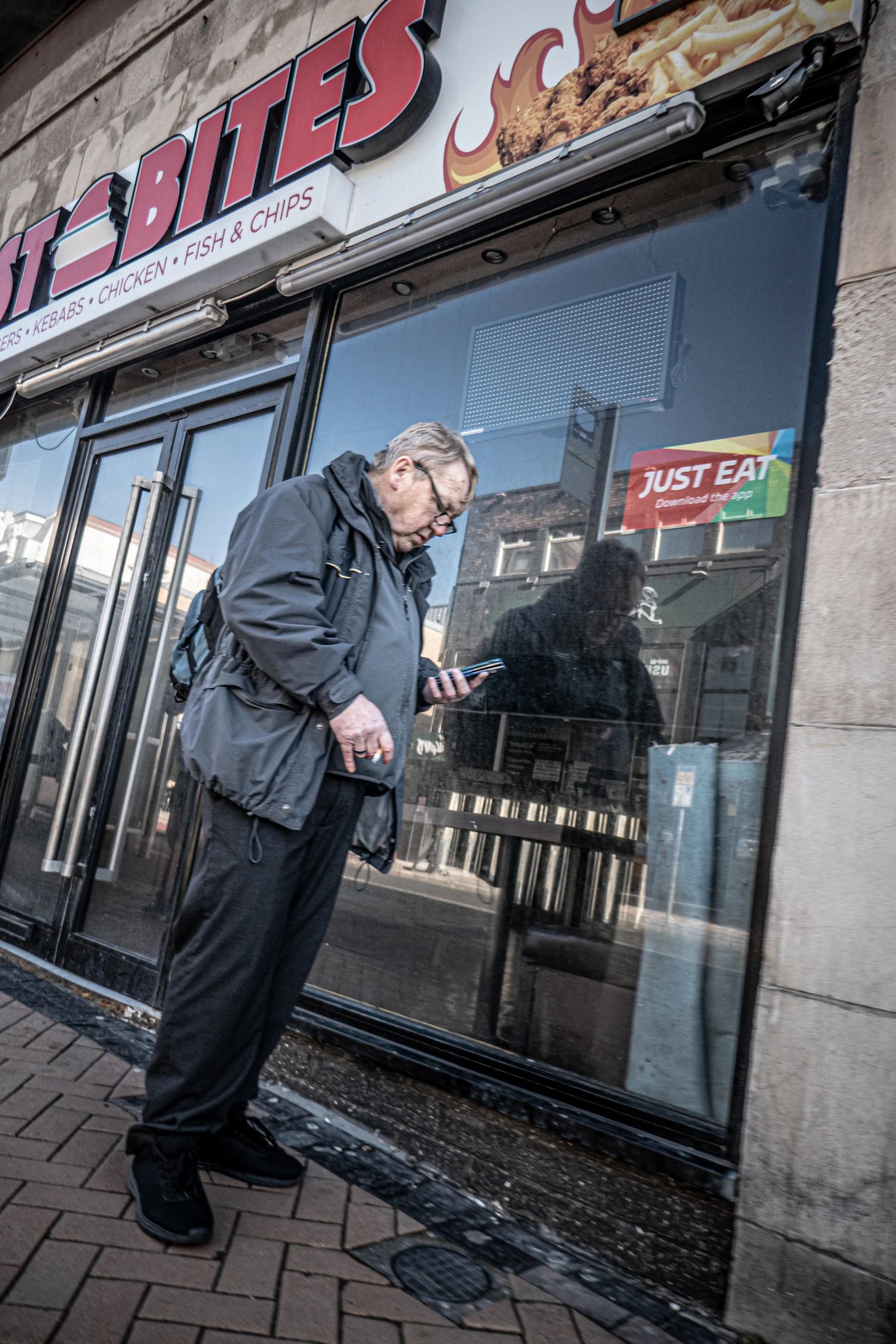 A man wearing glasses and a black jacket is standing outside a restaurant called Hot Bites, looking at his phone. The restaurant has a glass door and a sign advertising chicken, fish, and chips.