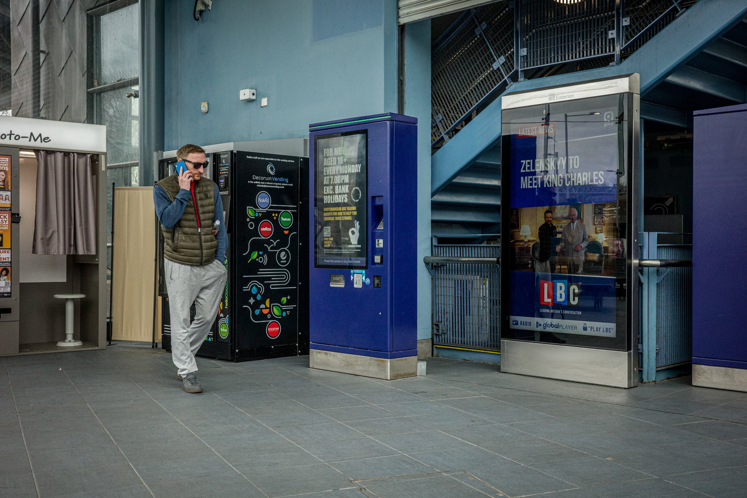A man standing outside a train station on the platform, talking on his cellphone and wearing sunglasses and a puffy vest. There are ticket machines and advertisements nearby, with a staircase leading up inside the station.