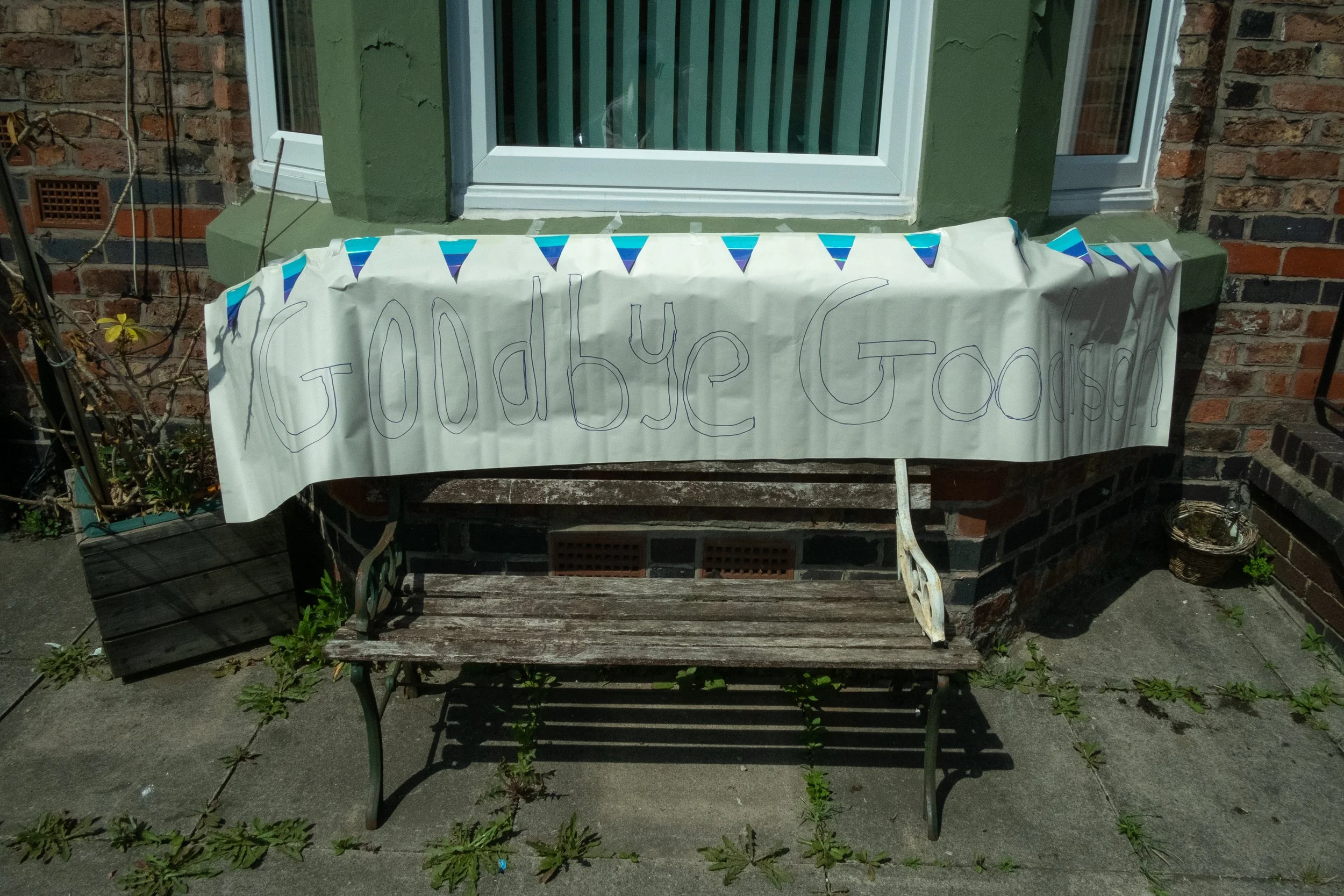 A handwritten sign on a paper banner hanging in front of a green window and brick wall reads 'Godbye Gouson' with colorful triangular decorations above.