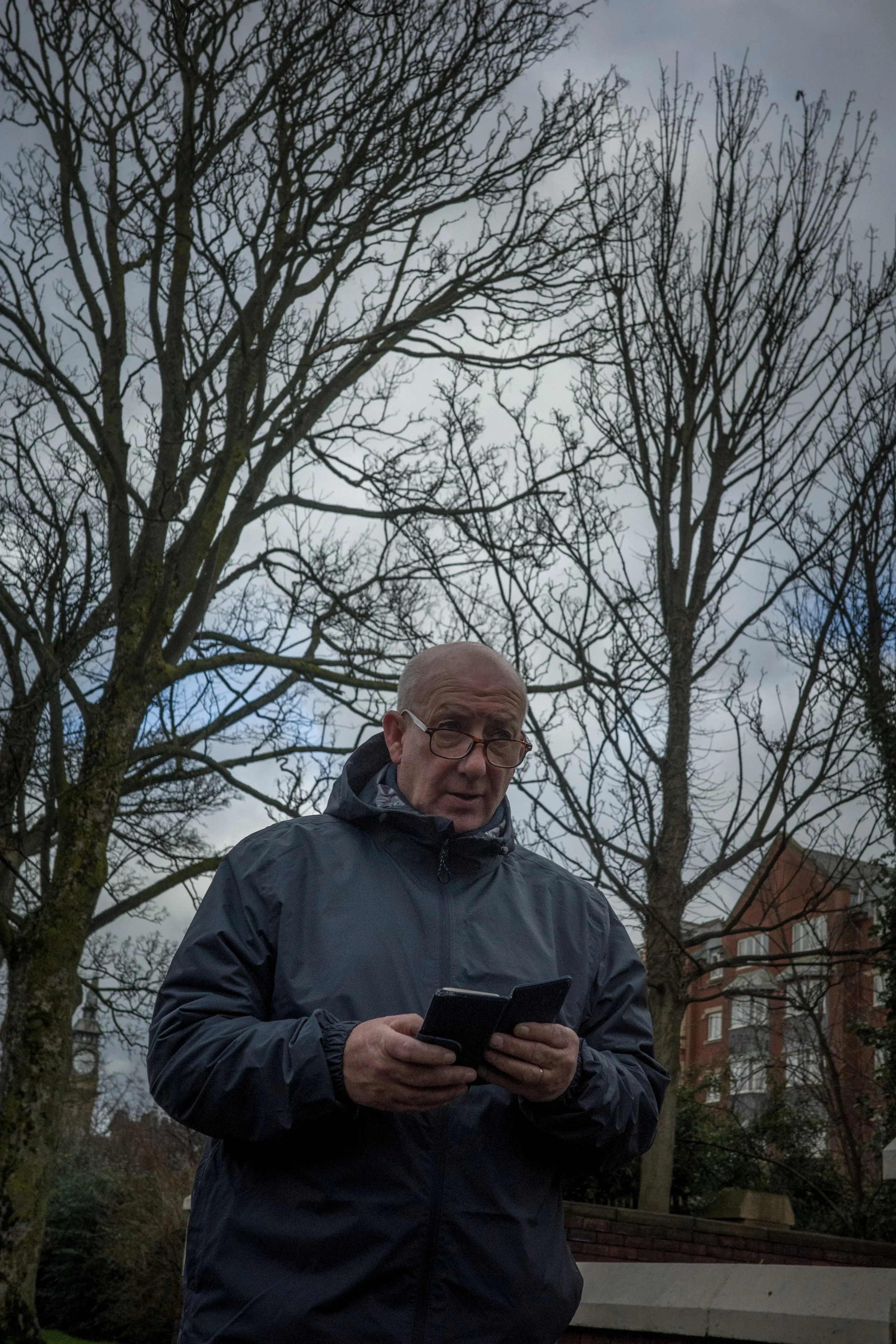 An elderly man wearing glasses and a dark jacket is outdoors during cloudy weather, looking at his phone with leafless trees and residential buildings in the background.