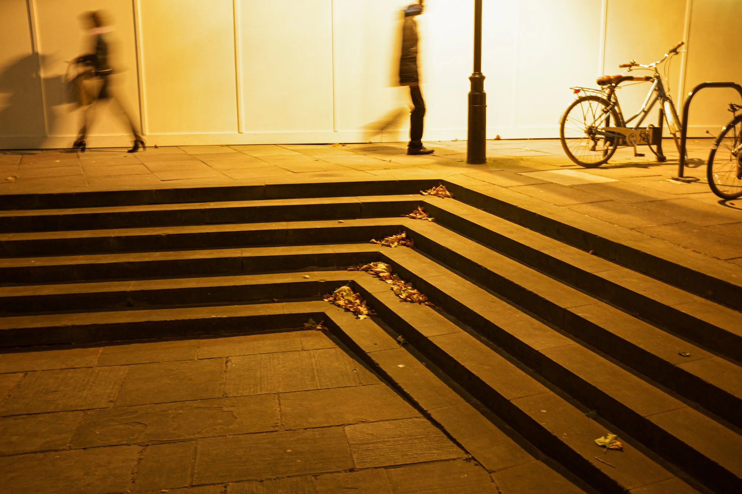 Street scene at night with stairs, fallen leaves, a lamp post, and bicycles parked near the sidewalk