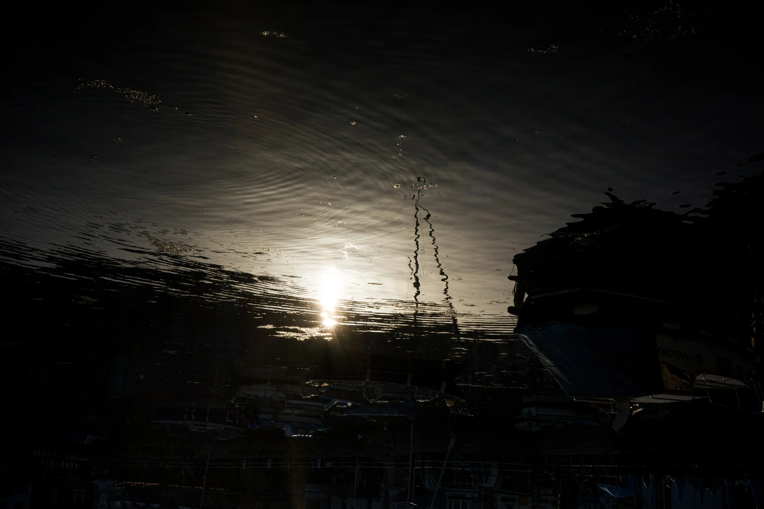 Sun reflecting on a calm body of water with ripples, with boats and masts visible upside down in the reflection