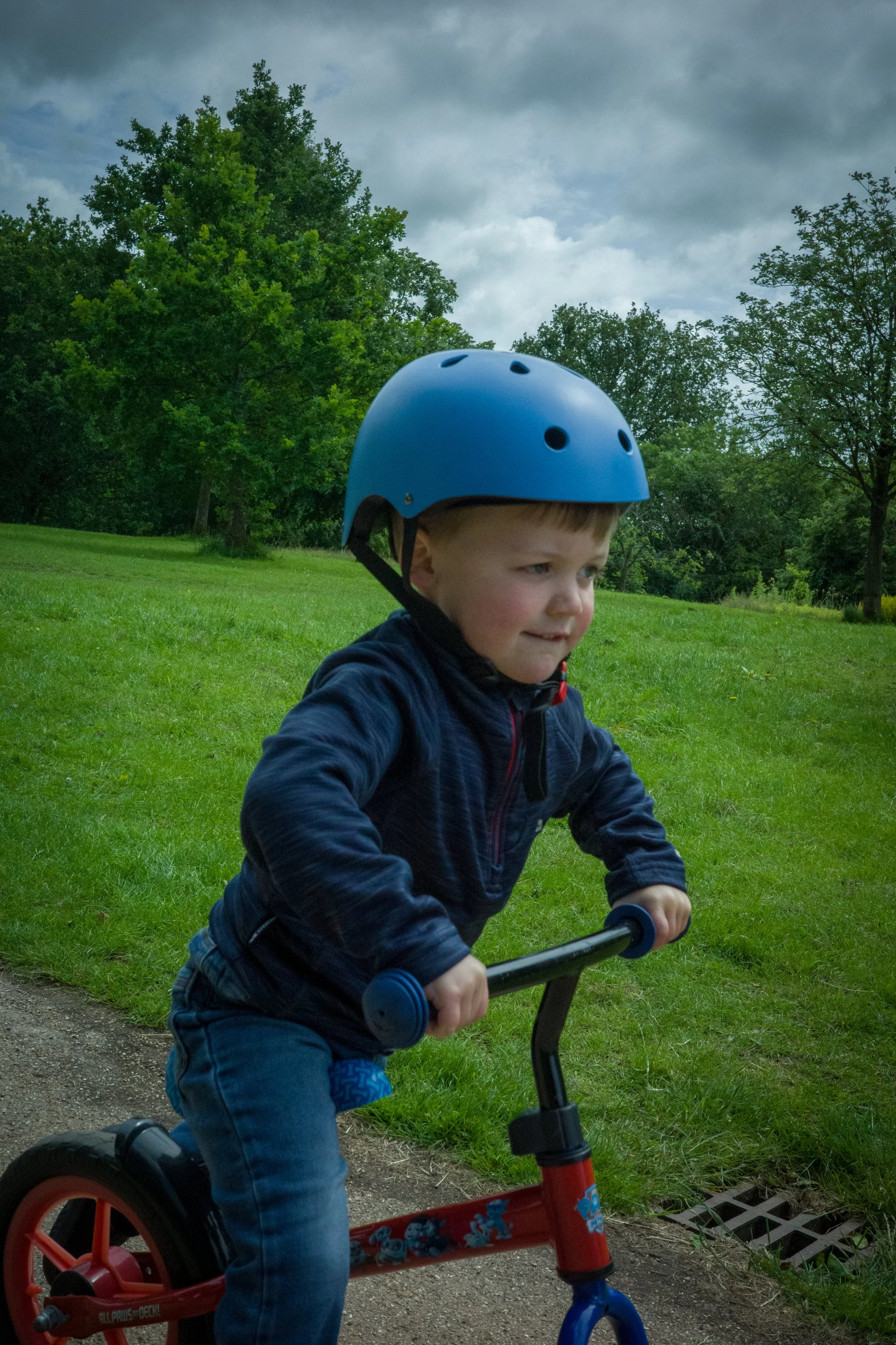 A young boy wearing a blue helmet and riding a red and blue balance bike on a park trail with green grass and trees, and cloudy sky.