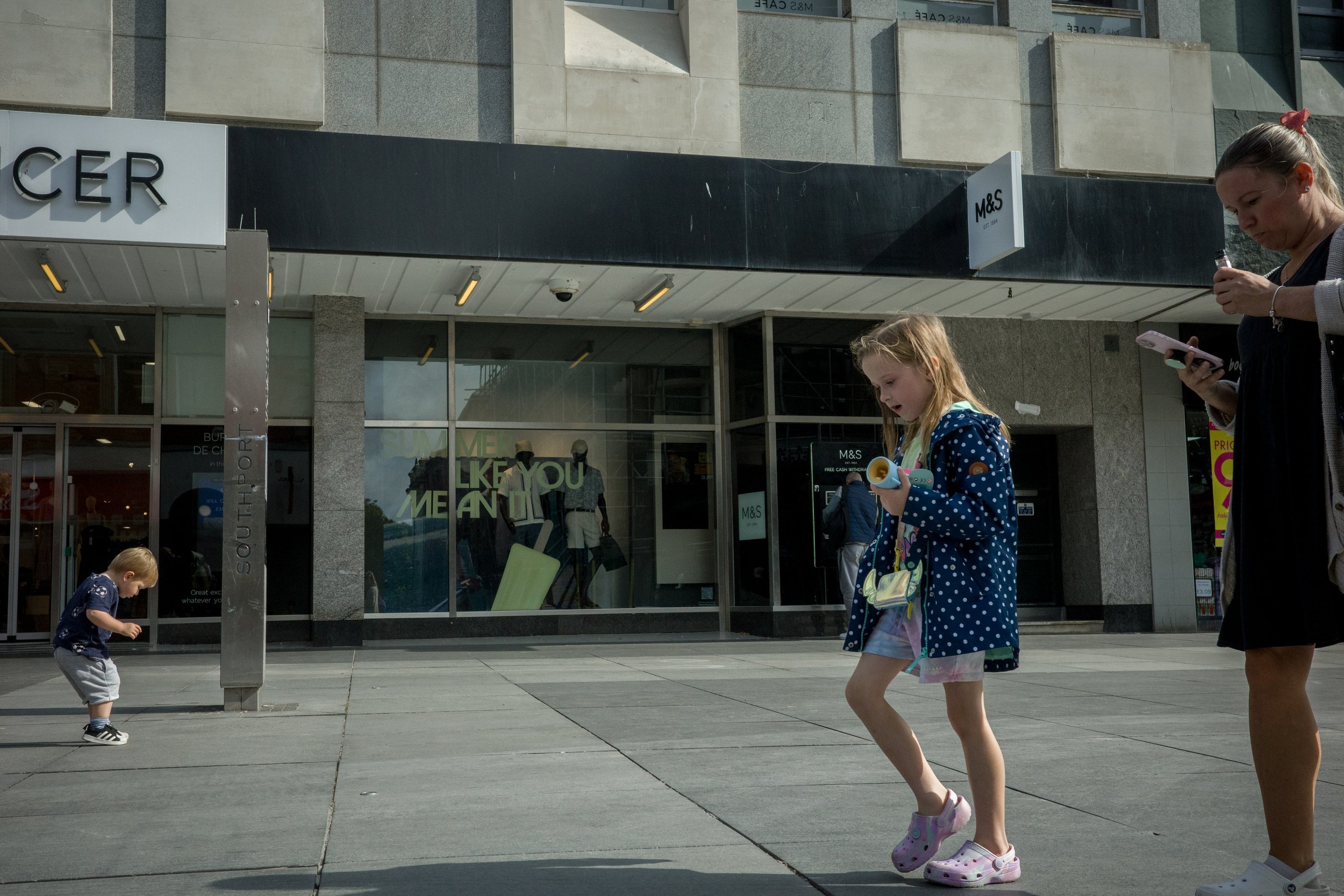 A street scene with a storefront for Marks & Spencer, three children playing on the sidewalk, and a woman standing nearby. One child is a girl in a blue polka-dot jacket and pink Crocs, and a boy in a navy shirt. The storefront has mannequins inside 