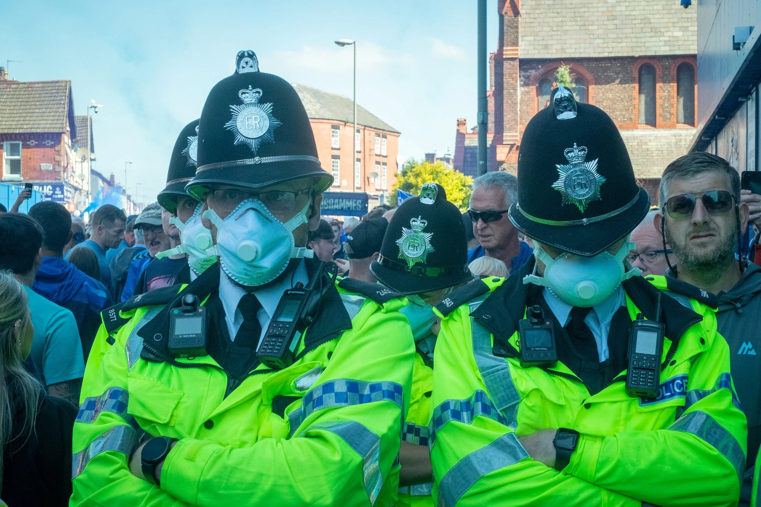 Two police officers wearing yellow high-visibility jackets, police hats, face masks, and radios stand with arms crossed at an outdoor event with a crowd of people behind them.