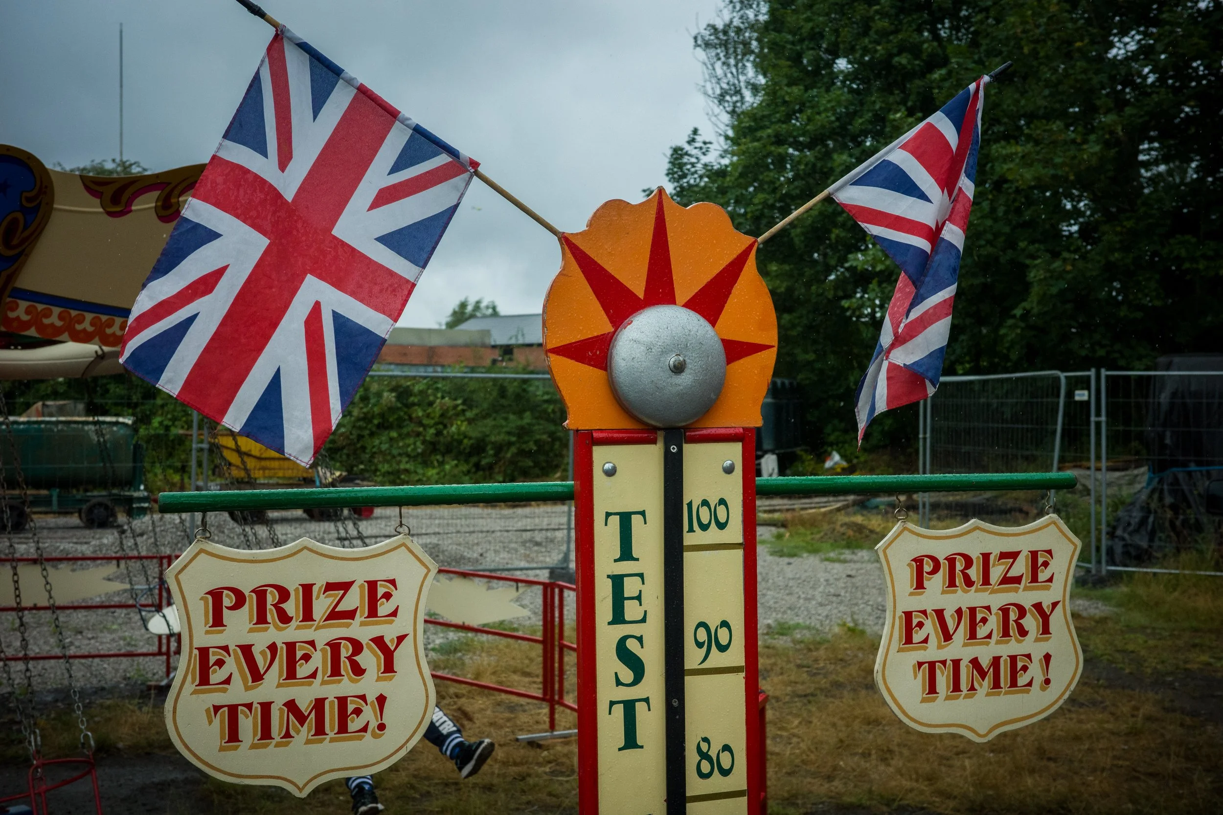 A carnival game booth featuring two British flags and a prize wheel with an orange center, surrounded by signs that read 'Prize Every Time!'