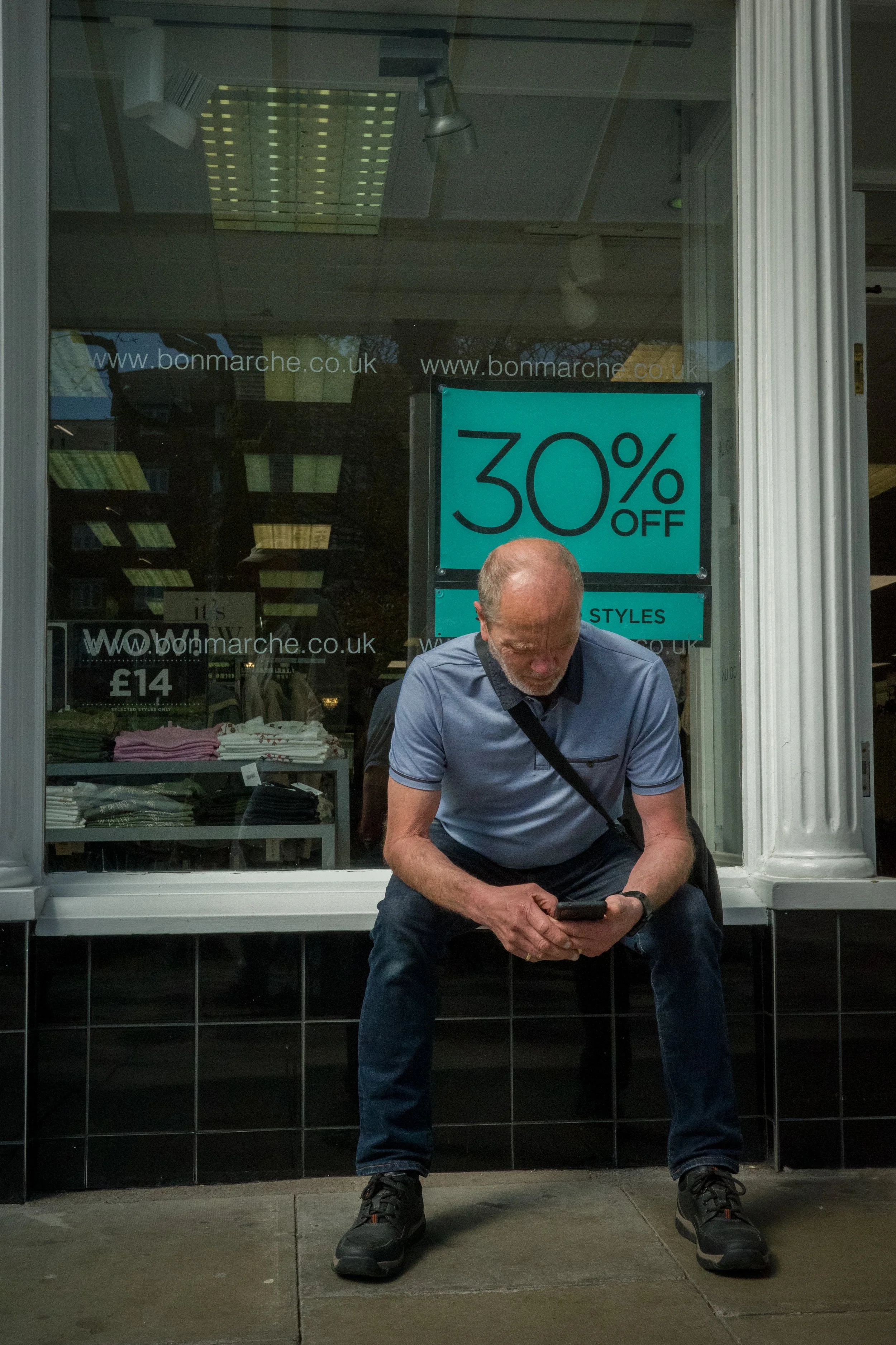 A man with gray hair sitting outside a store window, looking at his phone, wearing a light blue shirt, black jeans, and black sneakers, with a large turquoise promotional sign behind him indicating 30% off.