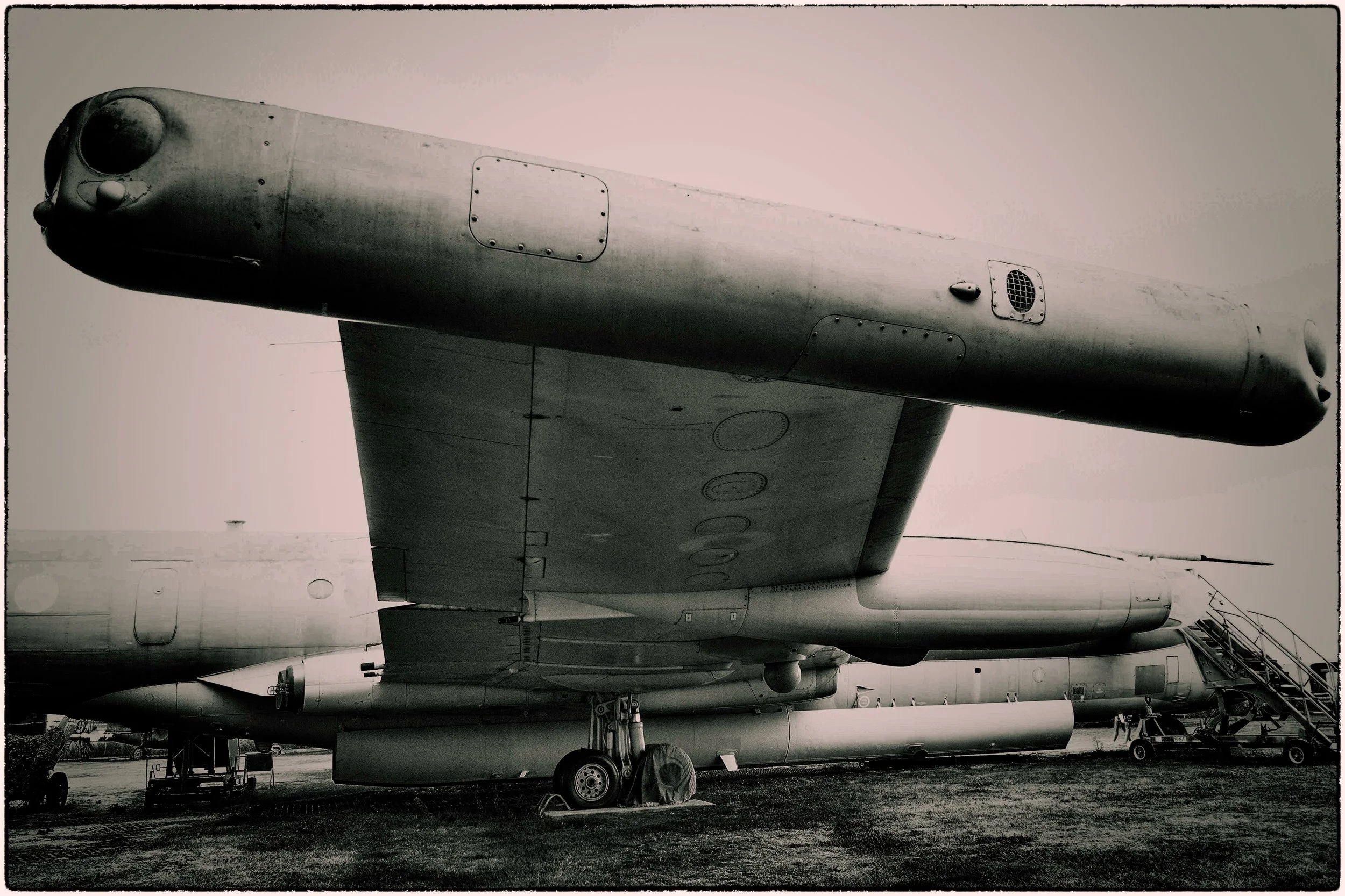 Black and white photo of a military jet aircraft on the ground, with various equipment and other aircraft nearby.