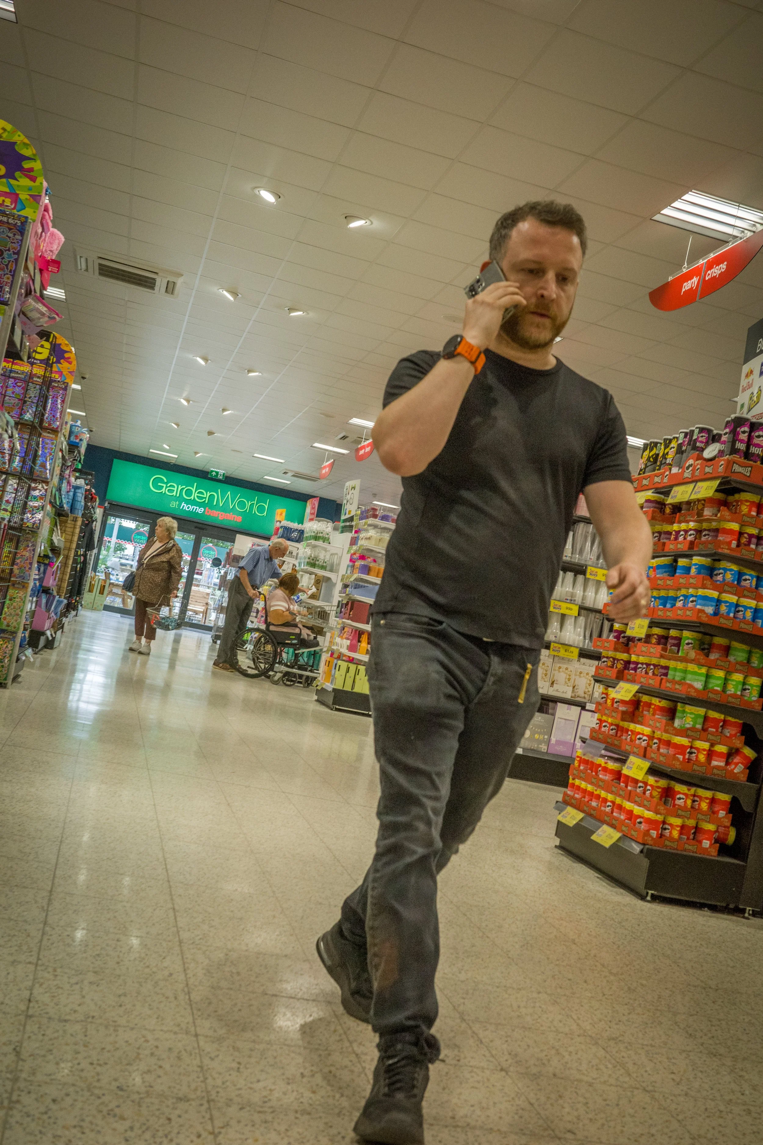 A man with a beard and dark hair walking through a store while talking on a cell phone. Shelves with various products line the aisle, and other shoppers are visible in the background.