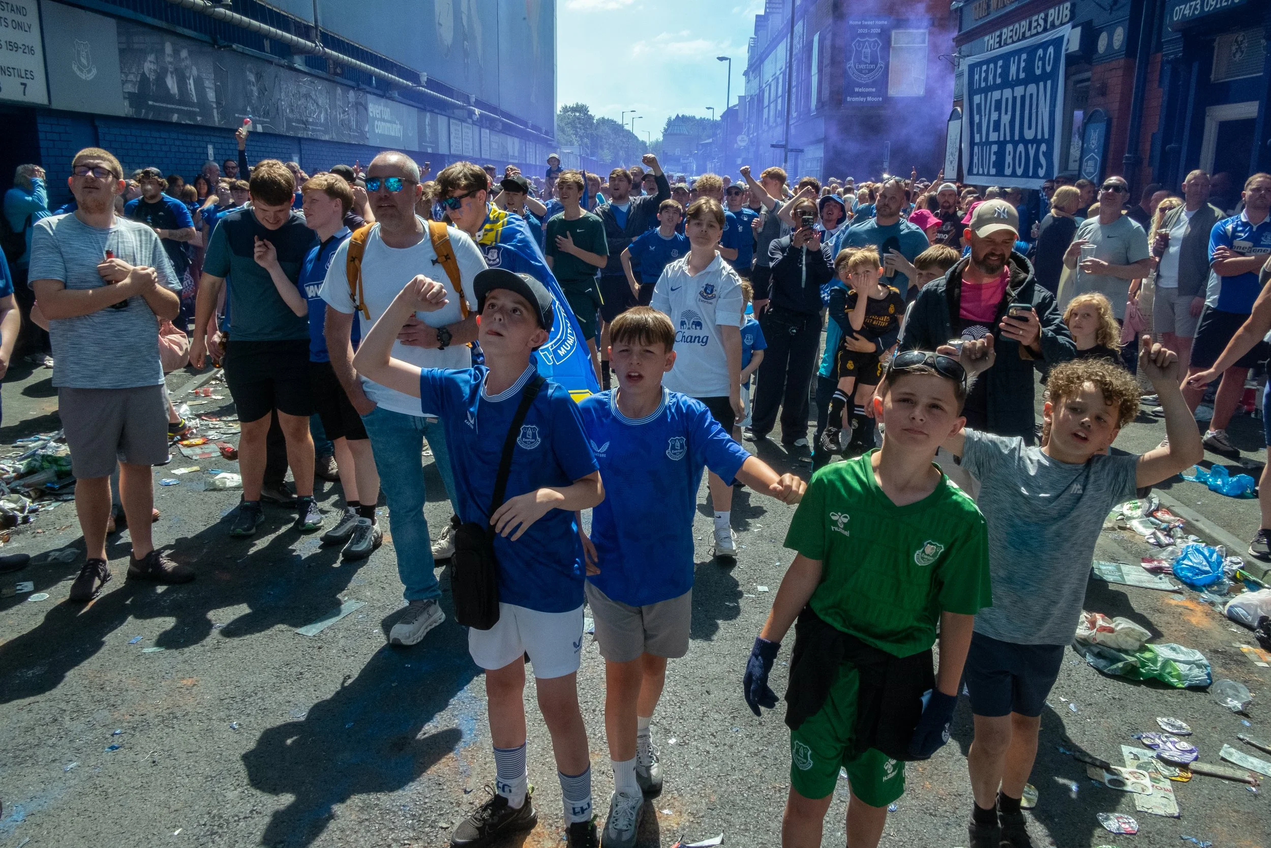 A crowd of soccer fans, including children and adults, gathered outside in a street, some wearing Everton jerseys, celebrating a match with some raising fists and holding drinks, amidst litter on the ground and signs supporting Everton.