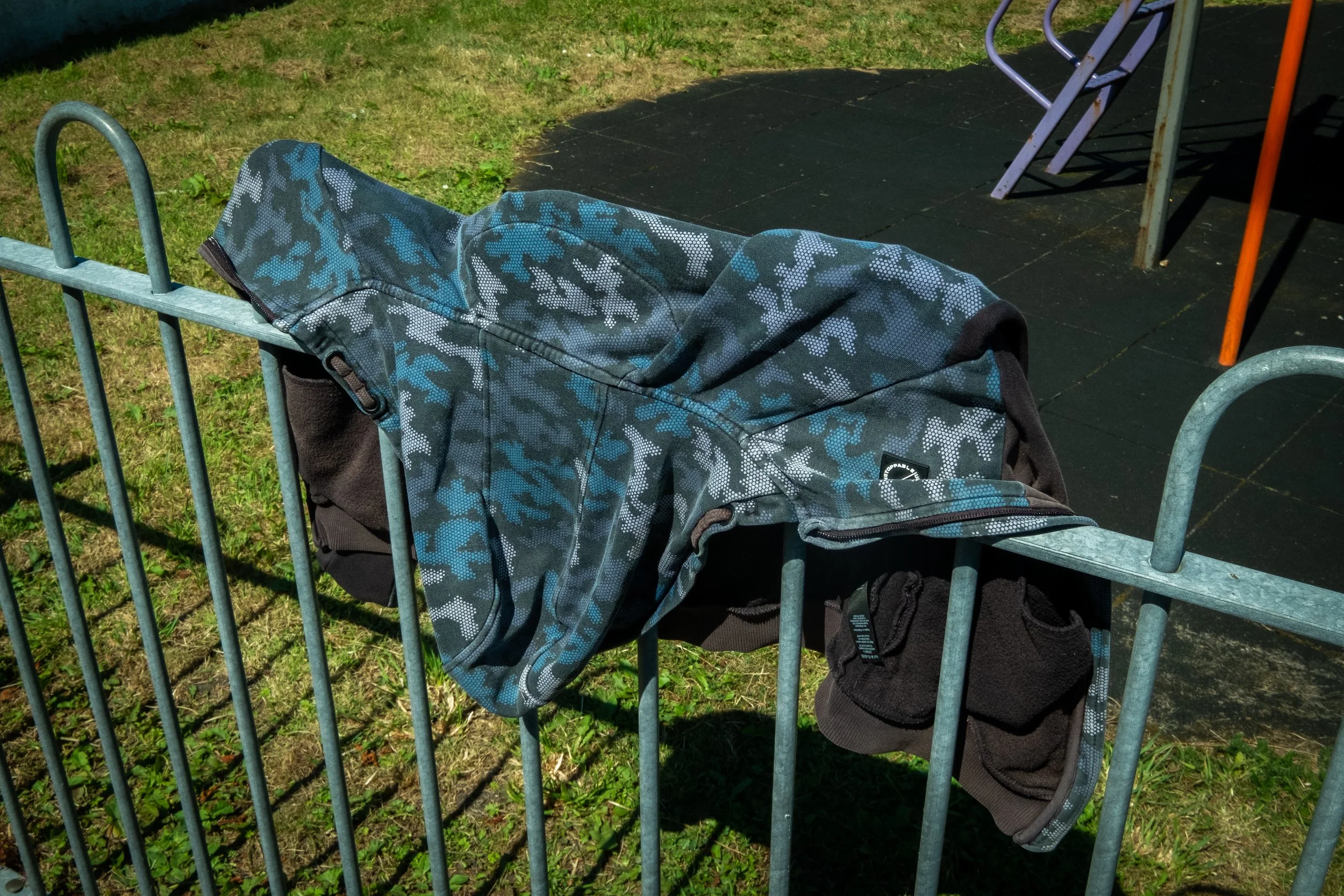A camouflage-patterned jacket draped over metal fence at playground with grass and equipment in background.