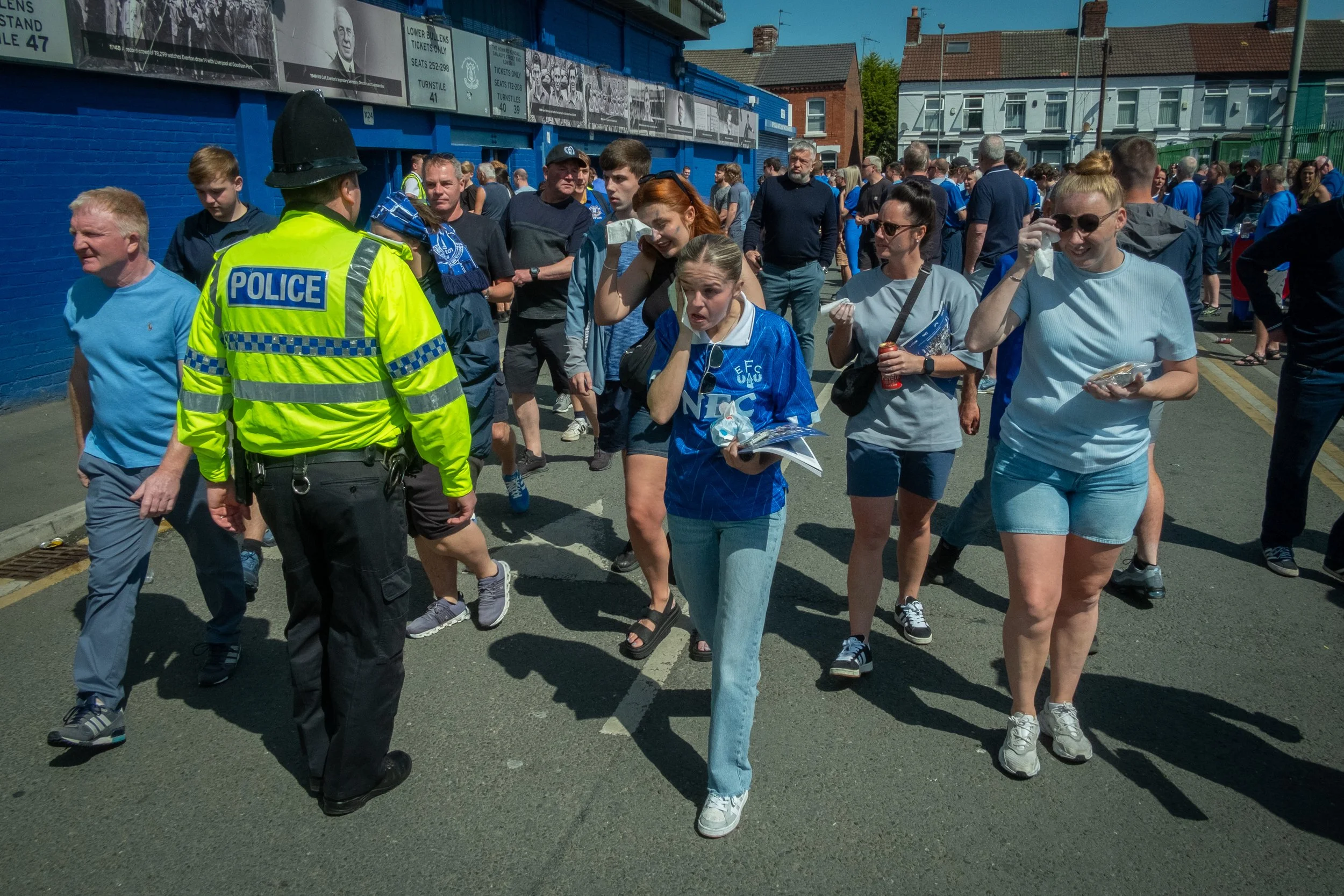 Crowd of people, including a police officer in a neon yellow jacket, walking outside in a sunny area