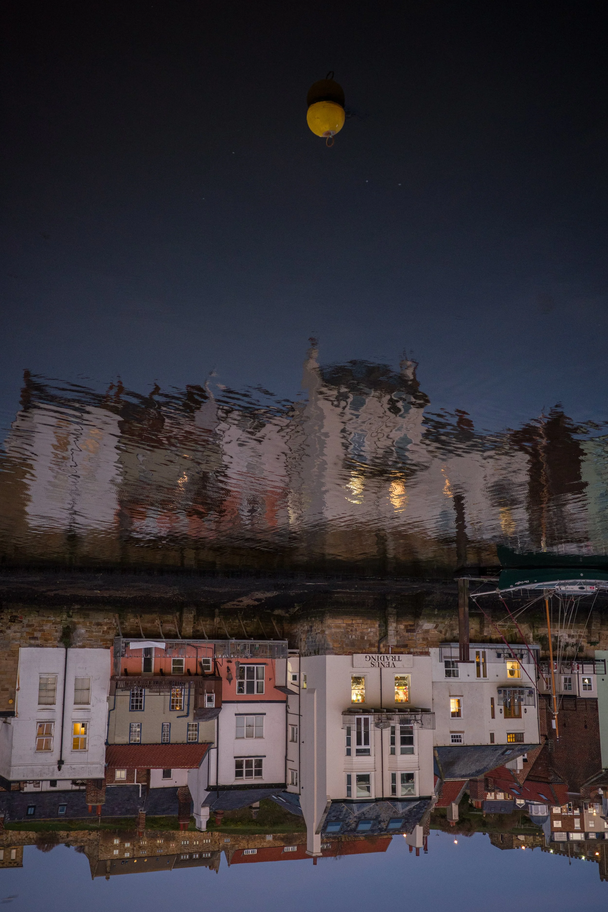 Upside-down reflection of buildings and a boat in water at dusk, with a yellow buoy floating above.