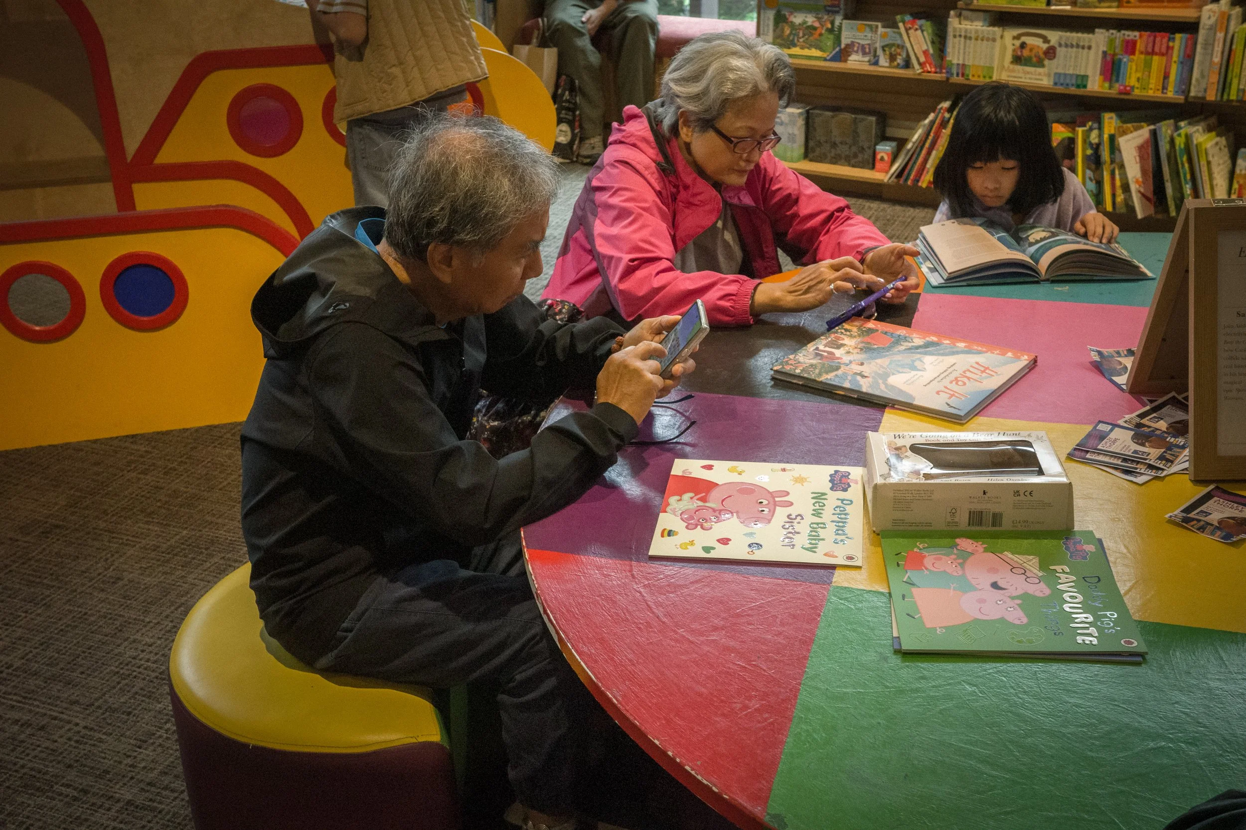 People sitting at a colorful table browsing and using their phones, with books and comics on display in the background.