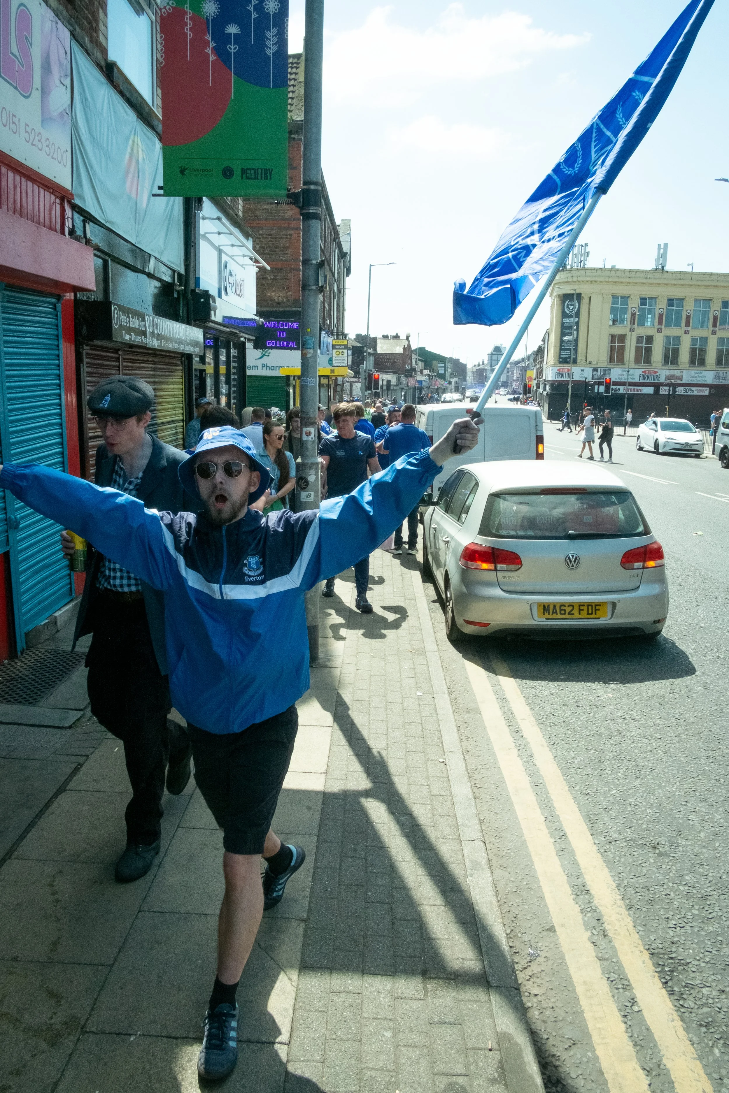 A man wearing sunglasses and a blue jacket is running on the sidewalk with a blue flag in his hand. Other people are walking behind him on a busy urban street.