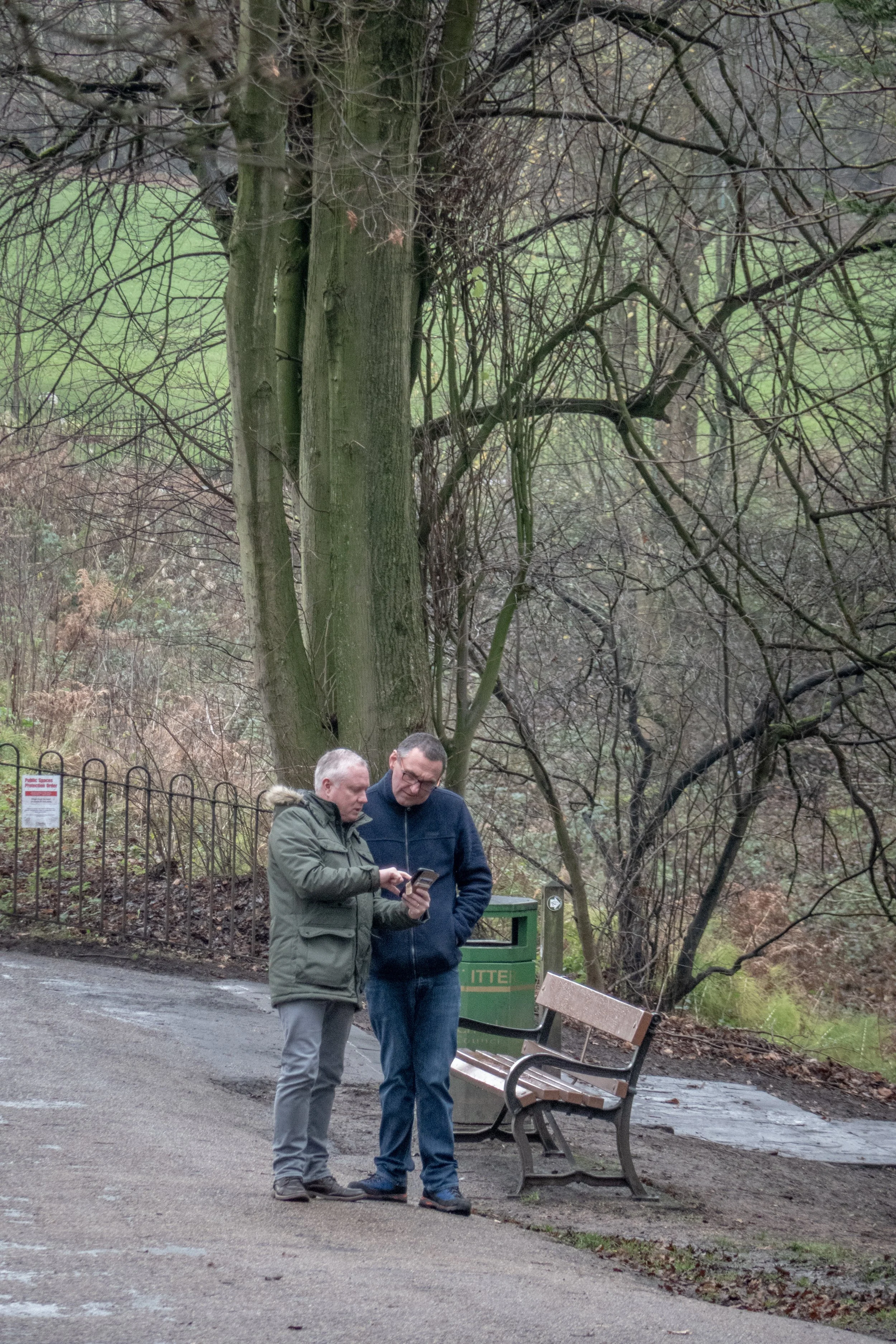 Two men are standing on a park walkway, looking at a smartphone together. One is wearing a green jacket, and the other is in a blue jacket. There is a bench, a green trash bin, and a fence with a sign behind them. Trees with bare branches and a grass