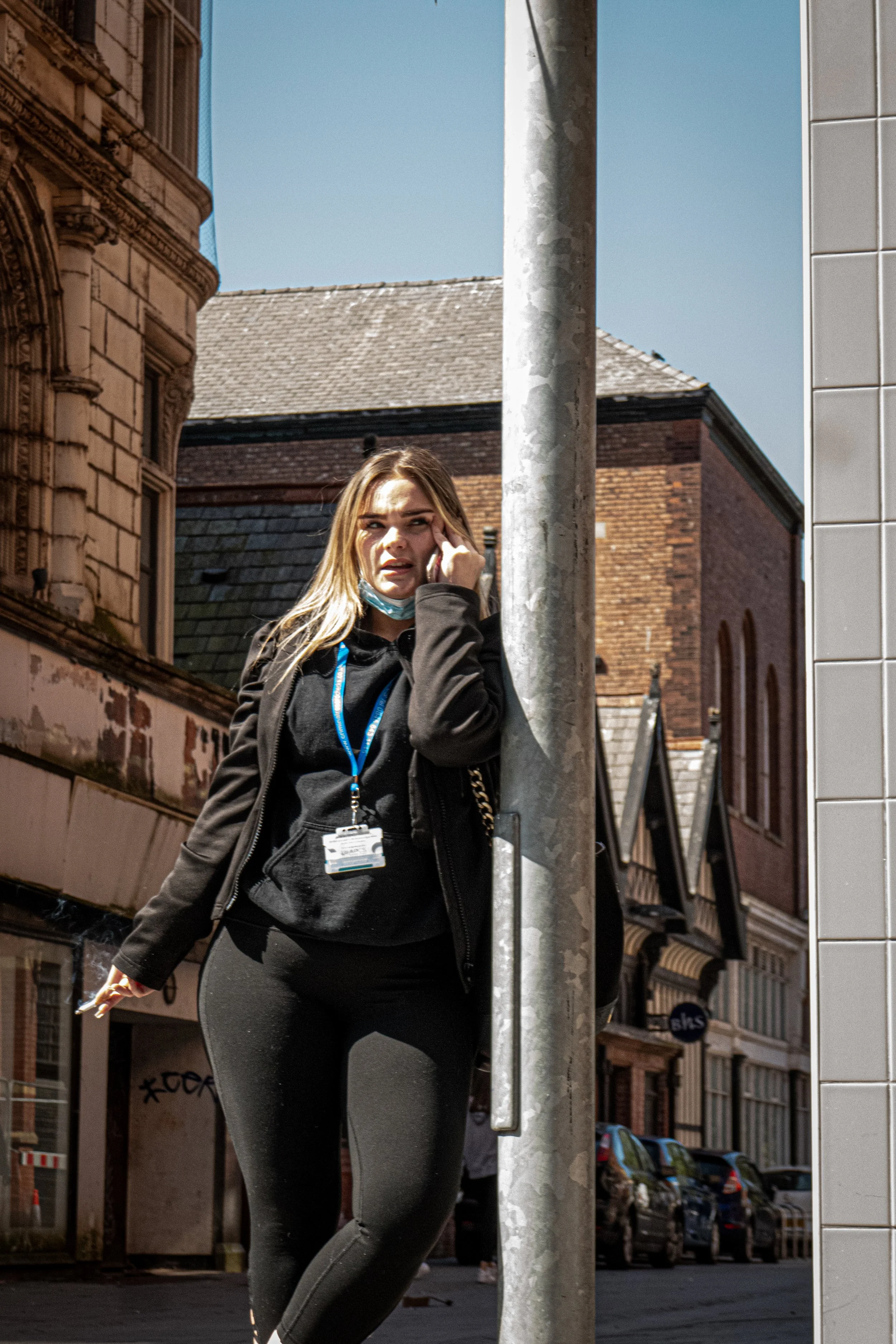 A young woman with long hair, wearing a black jacket and black leggings, stands outdoors talking on a cellphone, partially obscured by a metal pole, in an urban setting with buildings and cars in the background.