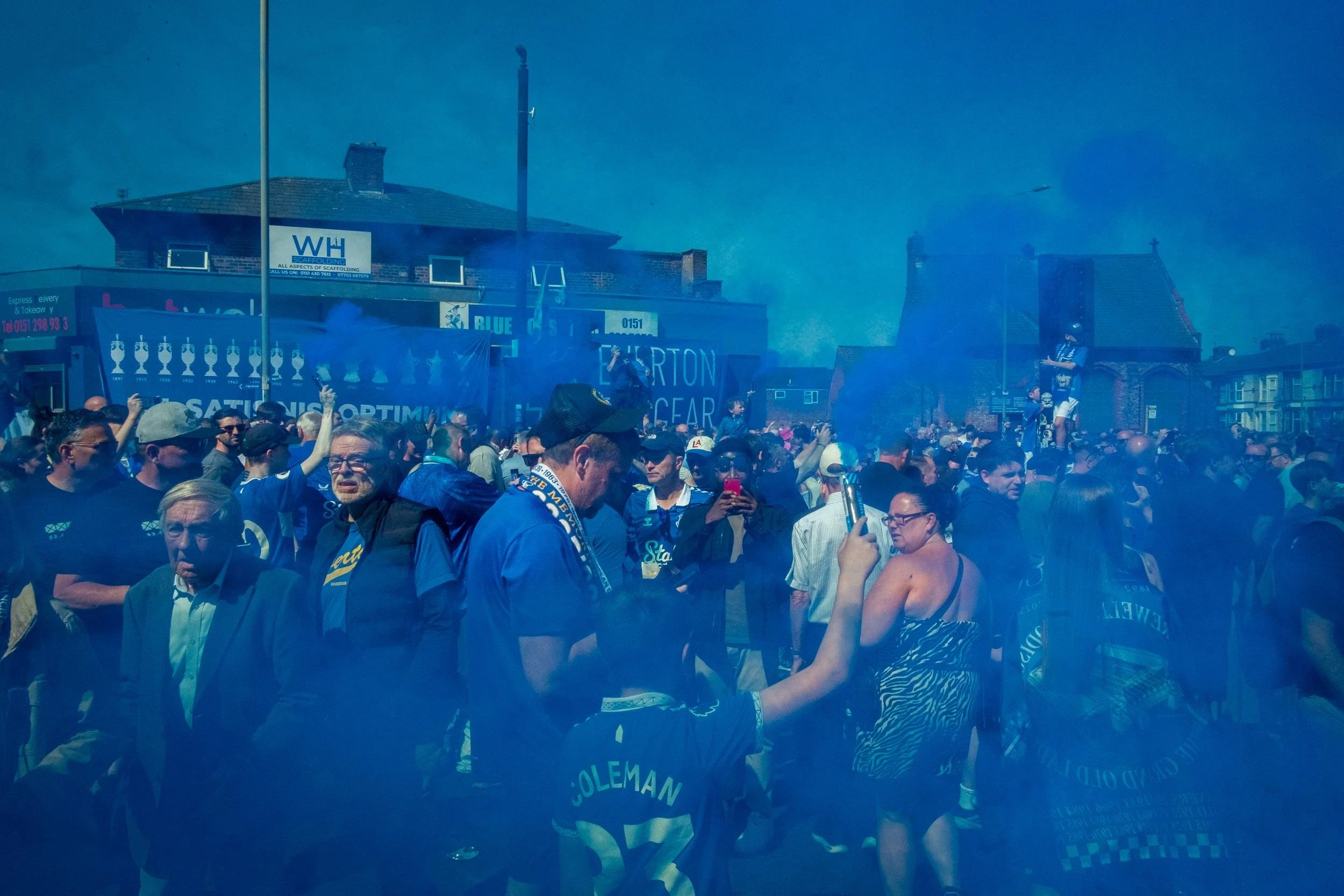 Crowd of people gathered outdoors, some wearing sports jerseys, with blue smoke in the air, celebrating or attending an event near a building with banners and signs.