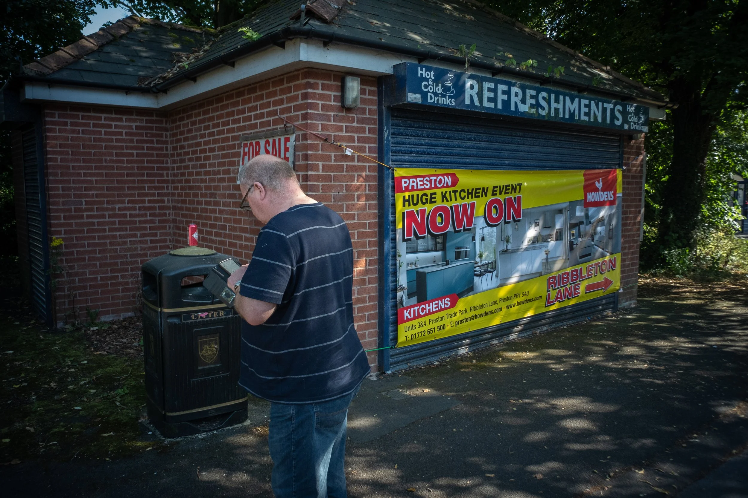 A man in a striped shirt using a public trash bin outside a small brick building with a sign for refreshments and a large yellow advertisement for a kitchen event