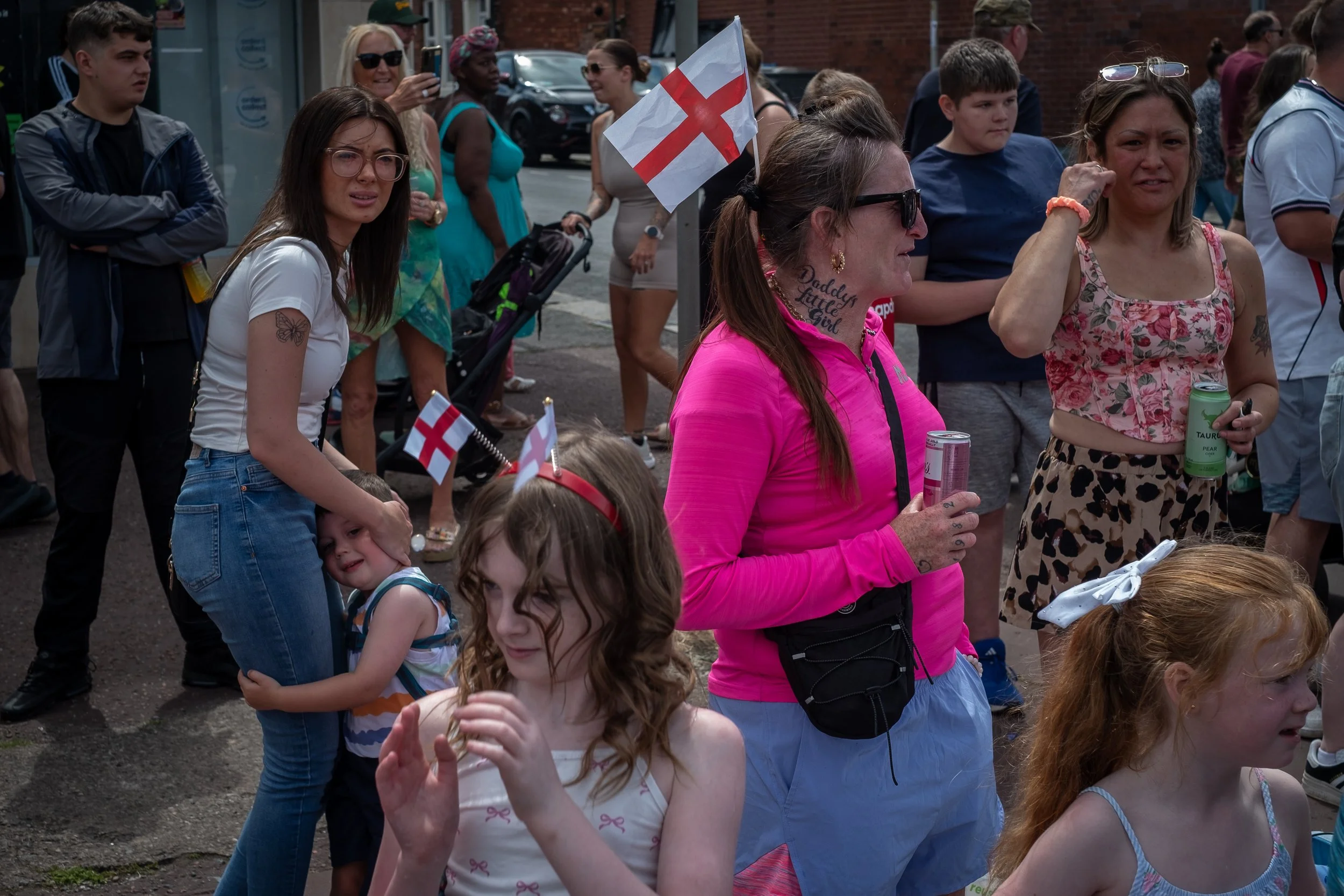 A group of people gathered outdoors, some holding small English flags, during a parade or public event. Women and children are visible, some talking or taking photos, with a woman in a pink top and sunglasses prominent in the foreground. Others are s