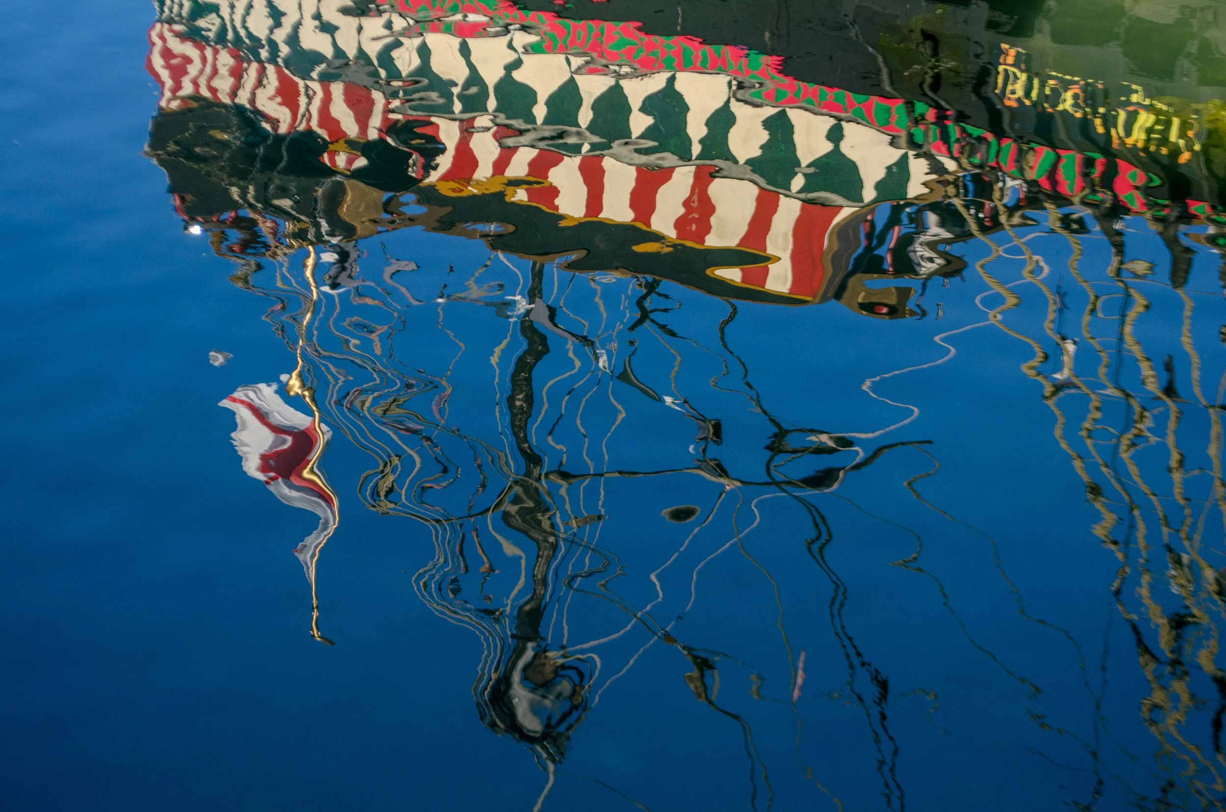 Reflected image of a boat with red, white, and green striped fabric and yellow lettering on the side, on the water's surface creating ripples and distorted reflections.