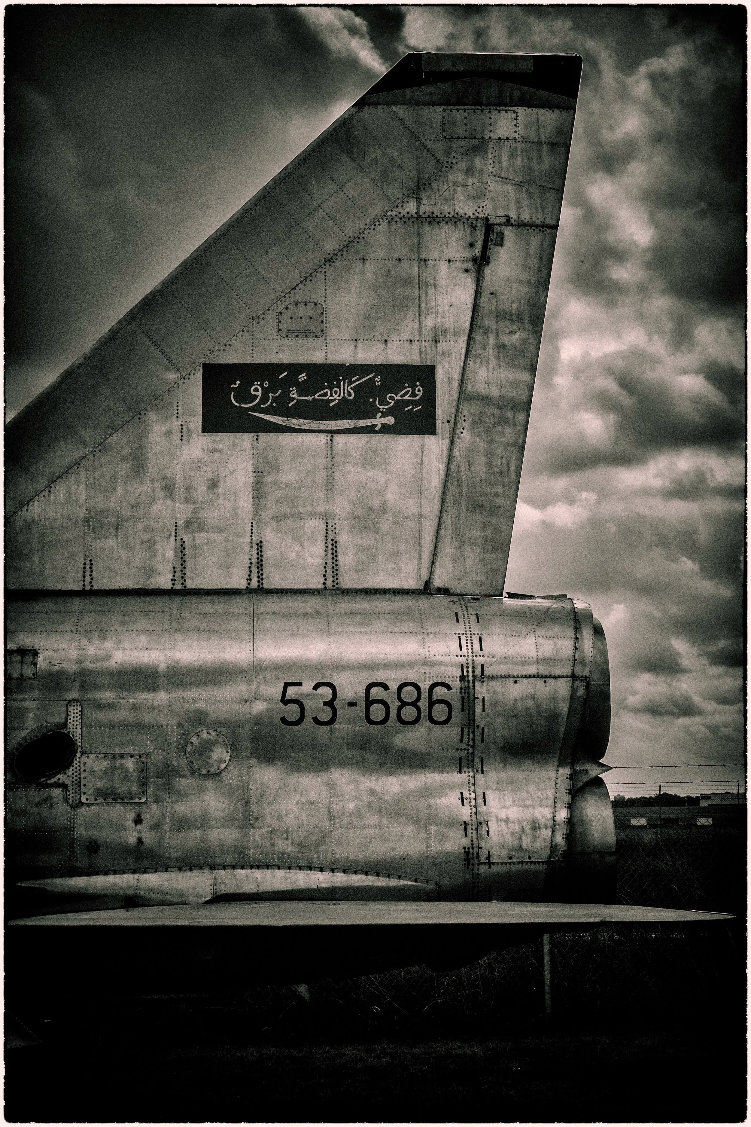 Close-up of the tail section of a military aircraft, showing the vertical stabilizer, part of the fuselage with serial number 53-686, and a black sign with Arabic text and a check mark. Dark clouds in the background.