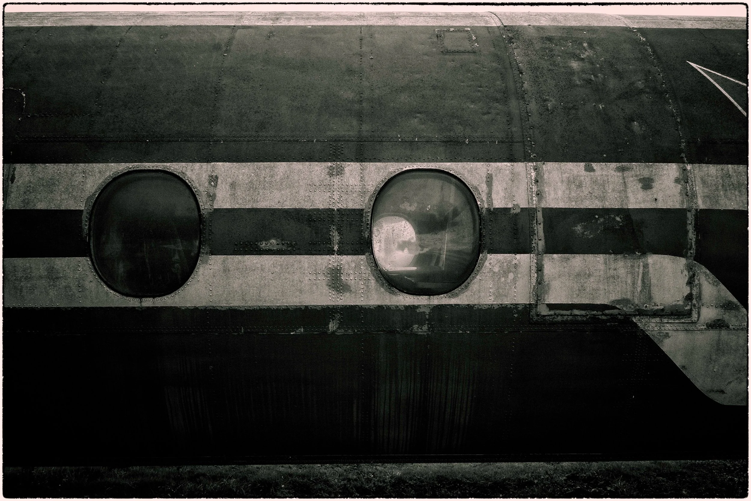 Close-up of a weathered metallic surface with two oval-shaped glass window panels, possibly part of an aircraft or industrial equipment, with rivets and visible wear.