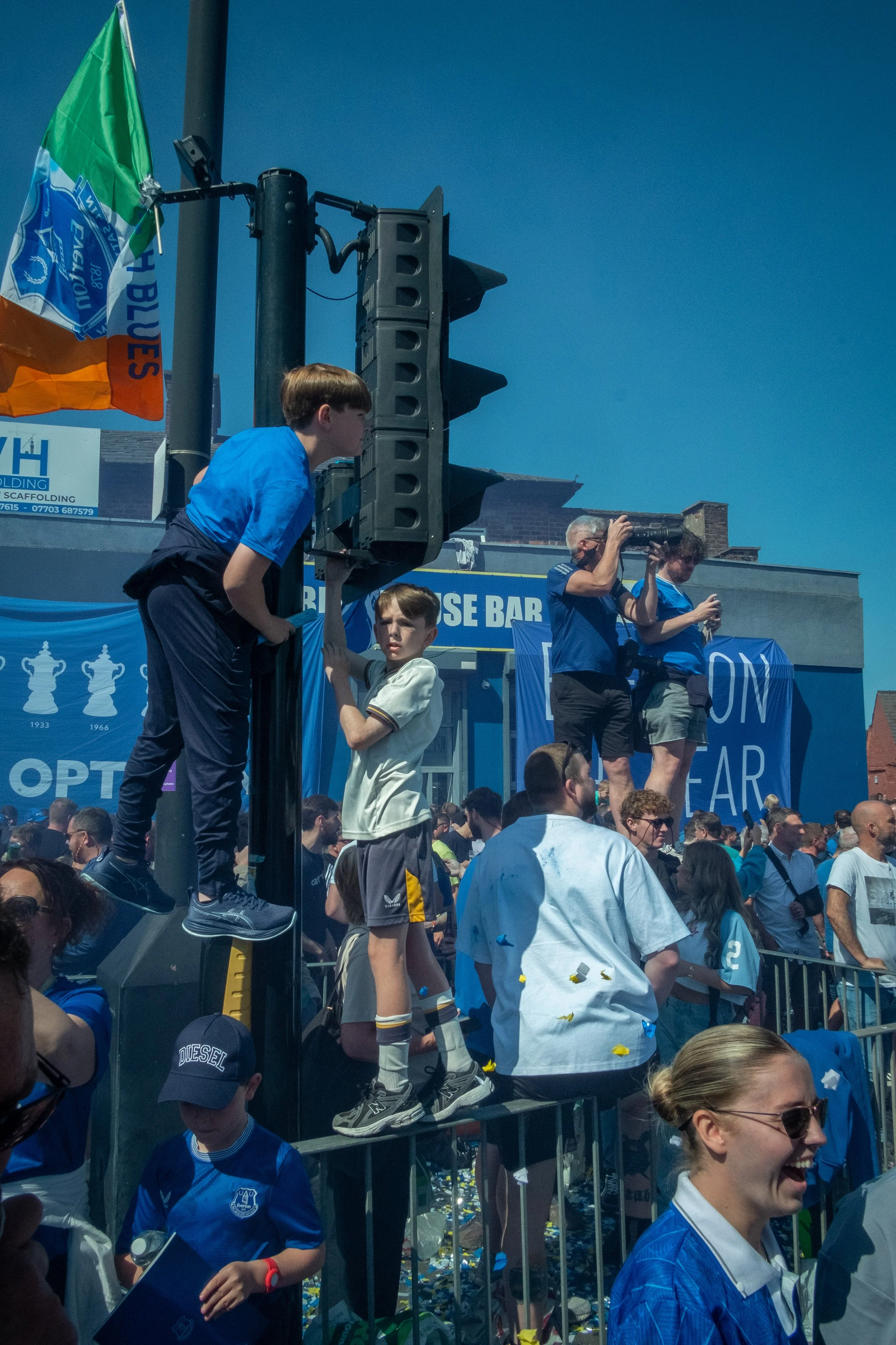 Crowd gathered at a public celebration or event, with people standing on barriers and congestion on the street. Some individuals are taking photographs, and there are blue banners and flags. The weather appears sunny and clear.