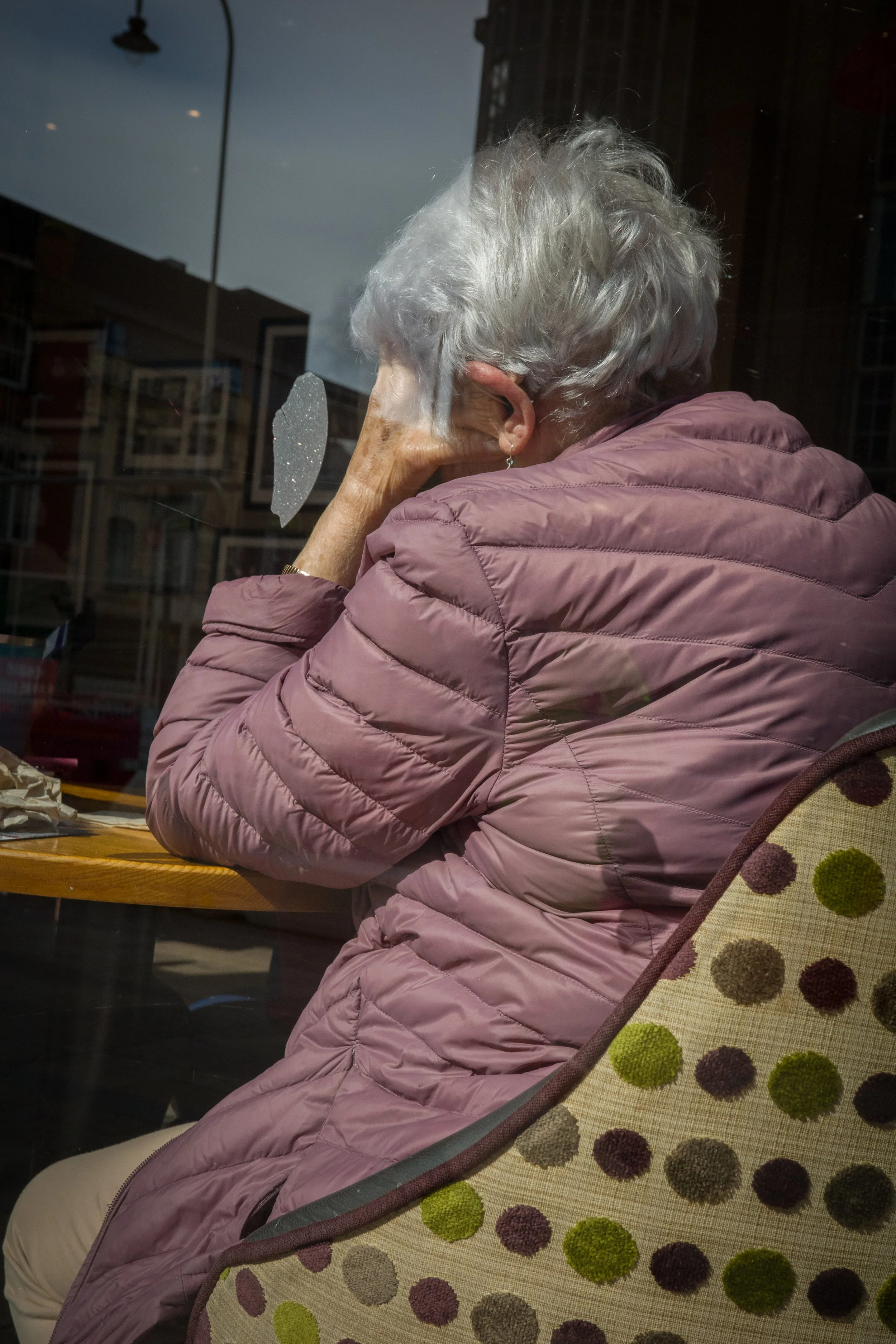 An elderly woman with white hair wearing a pink quilted jacket, sitting at a table inside a cafe, seen through a window with reflections and a broken sticker.
