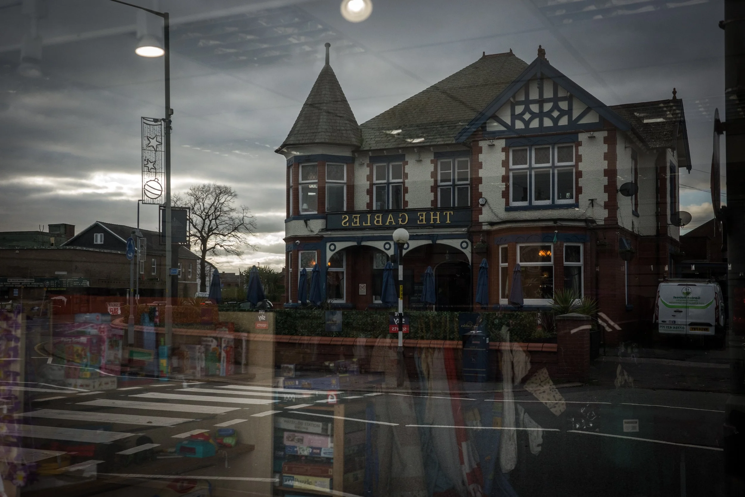A reflection of a house with a turret and a sign that reads 'The Gables' in a storefront window. The sky is cloudy, and there are trees and other buildings in the background.