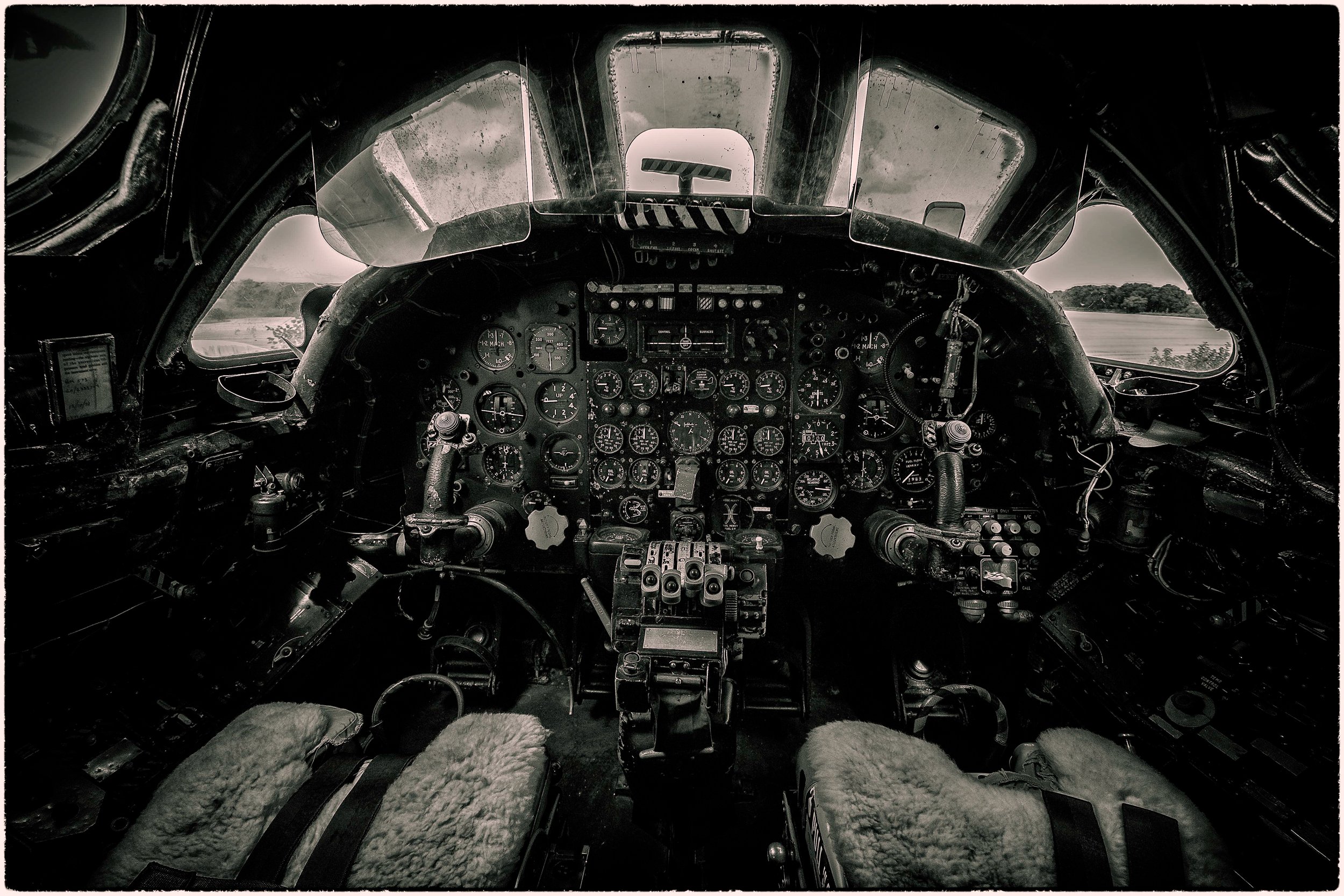 Interior of an airplane cockpit with numerous instruments, controls, and gauges, with worn seats covered in sheepskin, and a view of cloudy sky and landscape outside the windows.