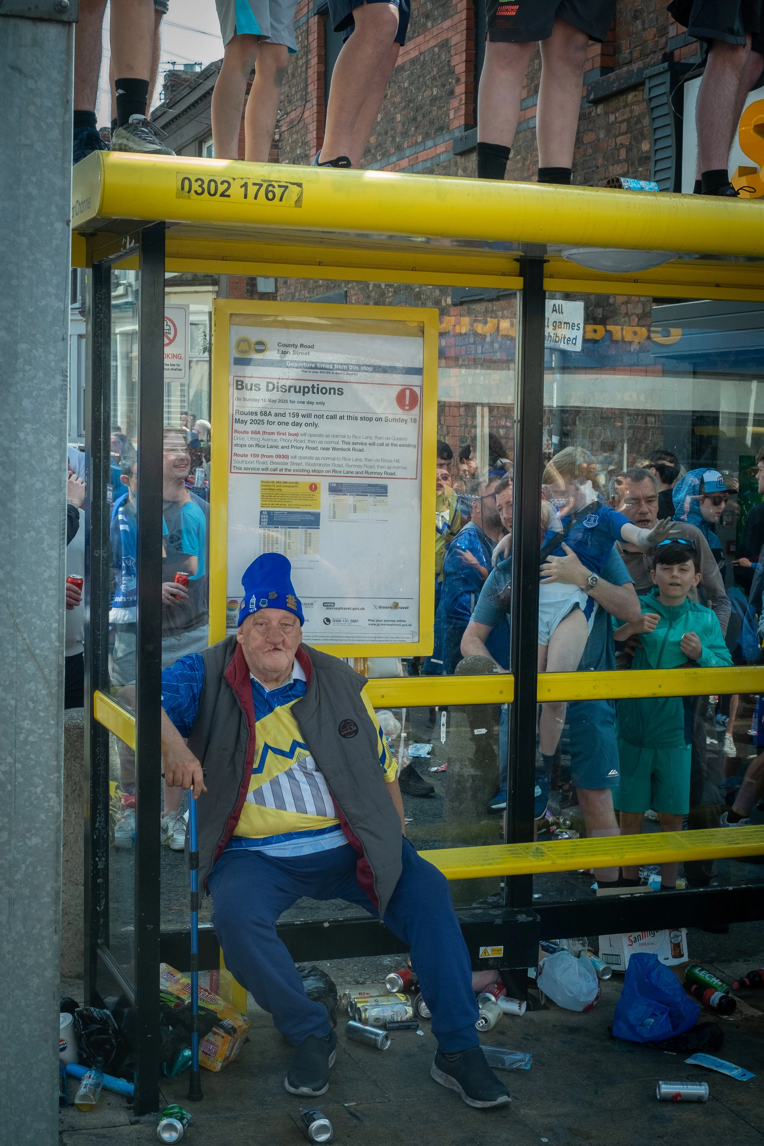 An elderly man sitting in a bus stop shelter surrounded by empty cans and bottles, with a crowd of people behind the glass, some lifting a young child, and a sign about bus disruptions visible.