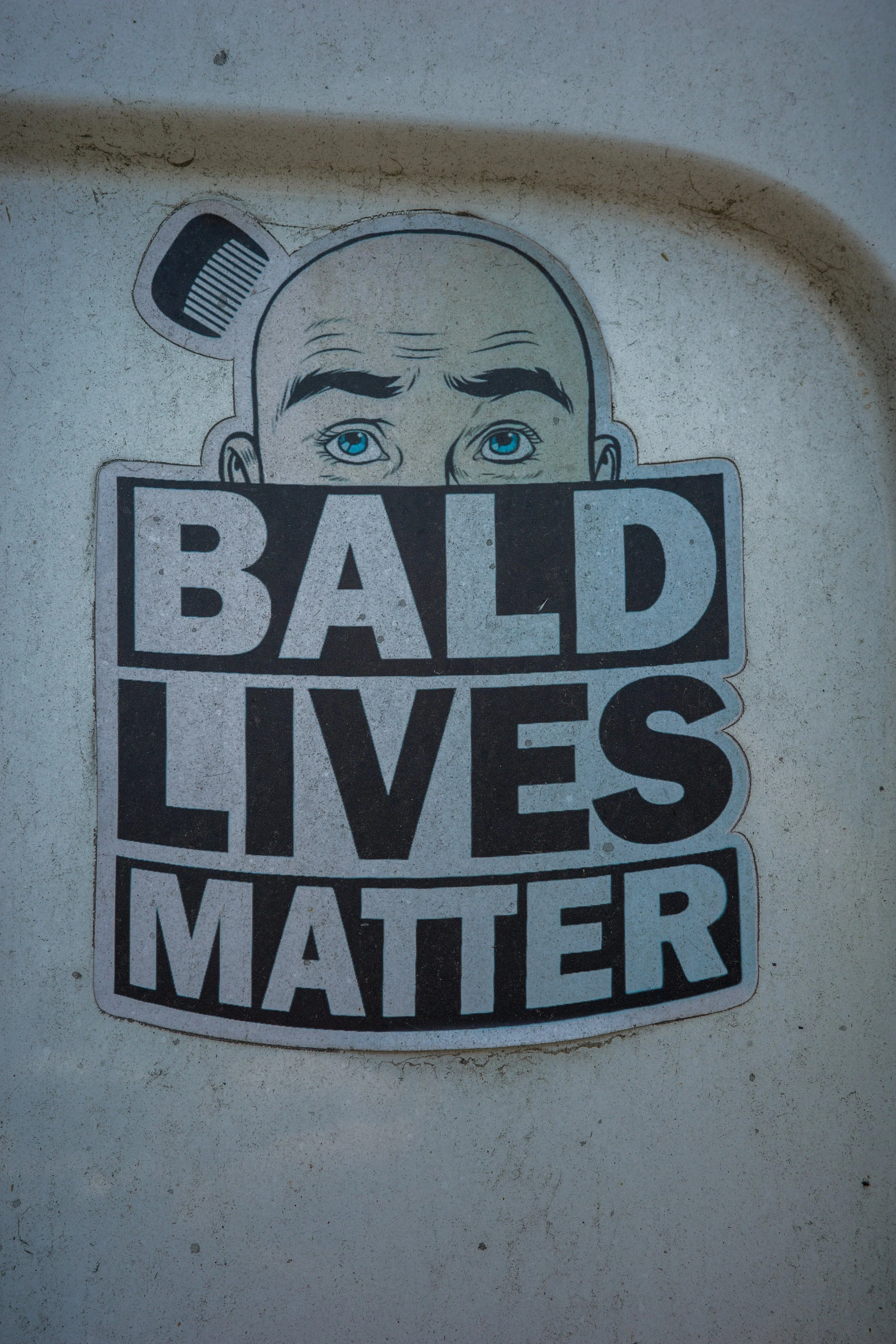 Sticker on a wall showing a cartoon face with bald head and blue eyes holding a sign that says, 'BALD LIVES MATTER'.