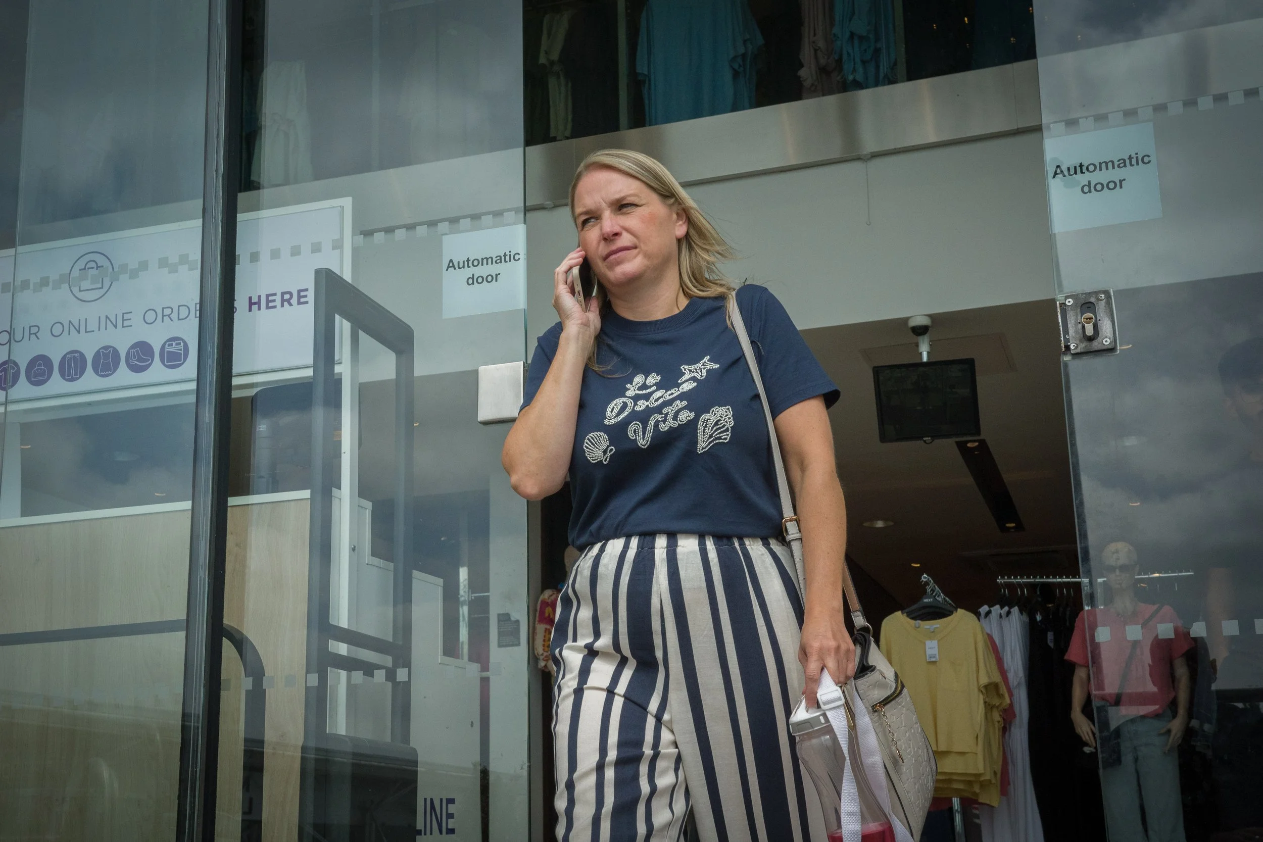 A woman with blonde hair talking on a cellphone, wearing a navy t-shirt with a seashell design and striped pants, standing in front of a glass door of a clothing store.