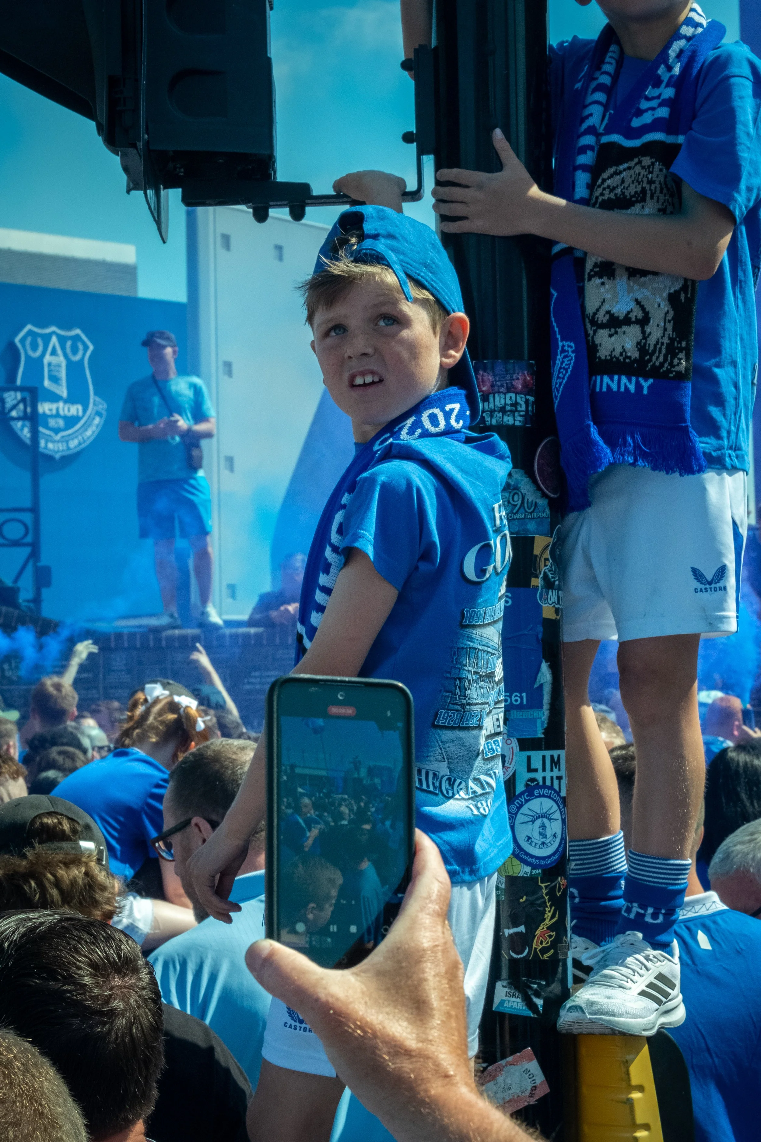 A young boy wearing a blue Everton football scarf and shirt stands on a platform at a large football gathering, with crowd around and fans in blue jerseys. A person in the background is holding a phone to record the scene.