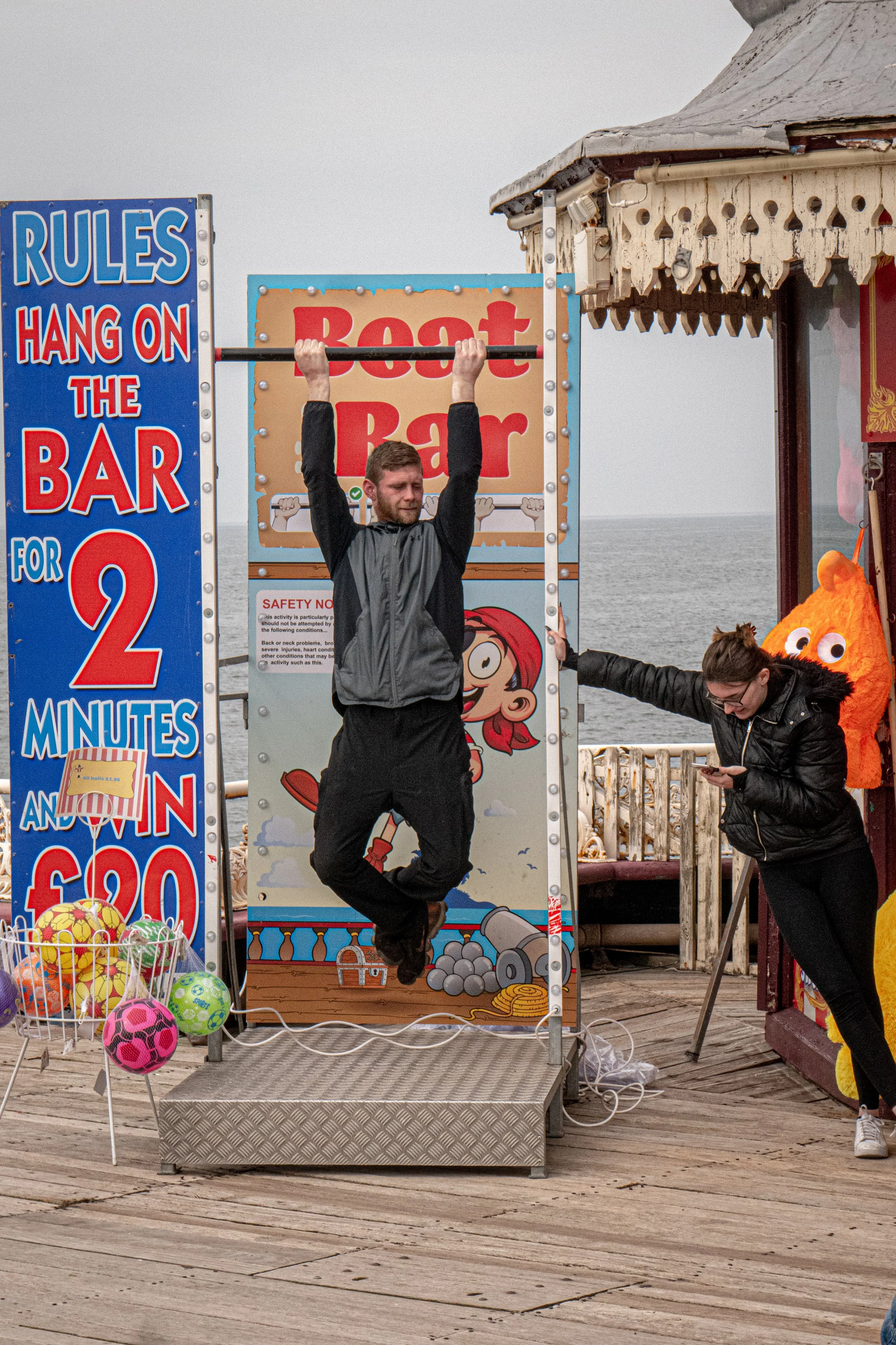 A man hanging upside down from a bar in a fair game of strength on a wooden boardwalk near the ocean, with a woman standing nearby, a colorful mascot costume, and signs advertising the game.