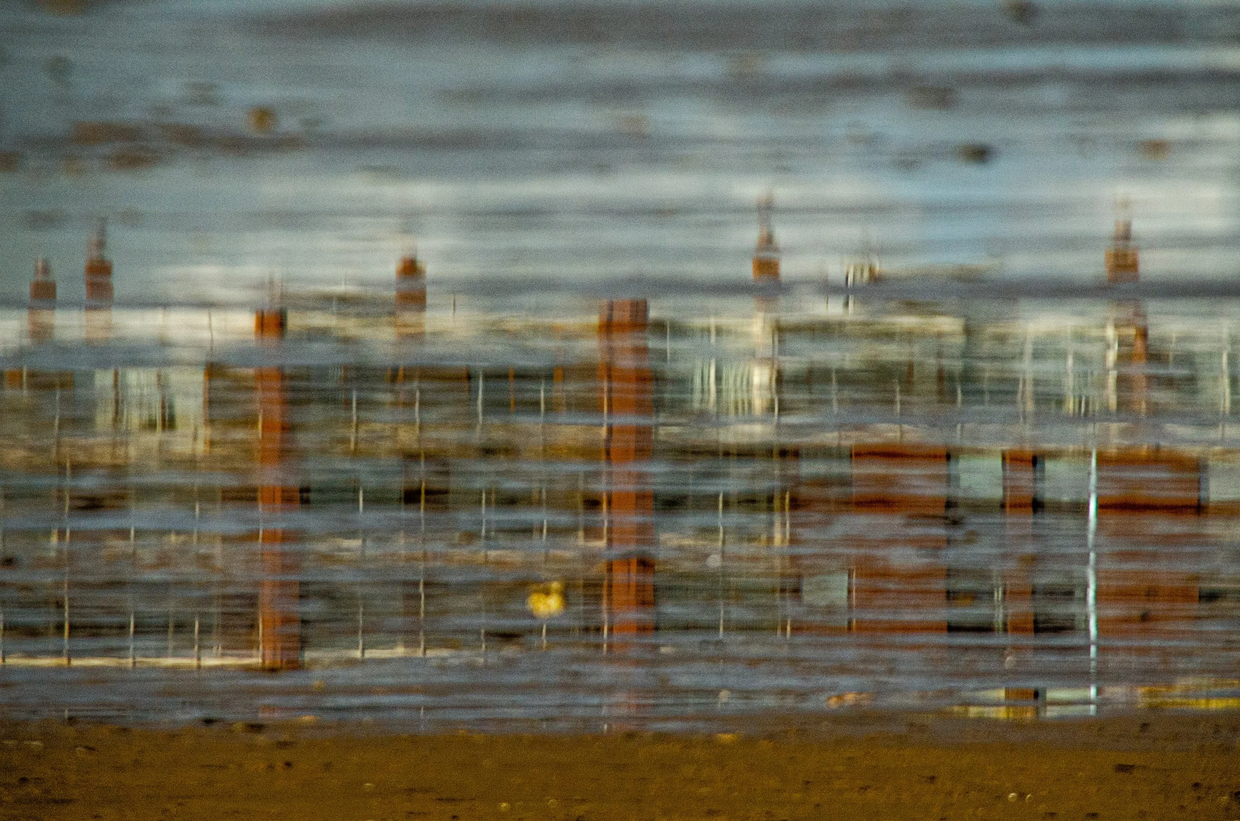 A blurred view of a construction site behind a chain-link fence near a body of water, with some buildings visible in the background.